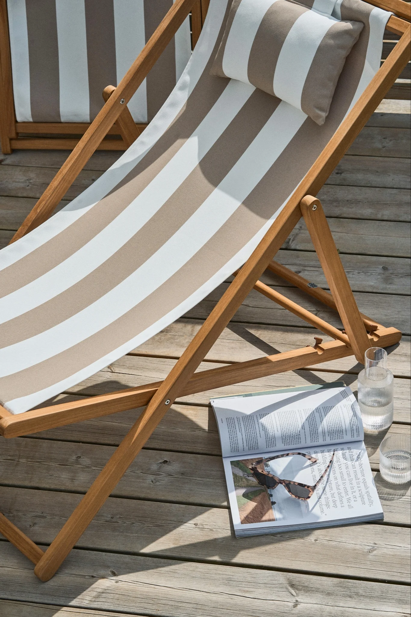 Ein hölzerner Liegestuhl mit gestreifter Auflage und Kissen, einer Zeitschrift mit Sonnenbrille und einem Glas Wasser auf einer Holzterrasse.