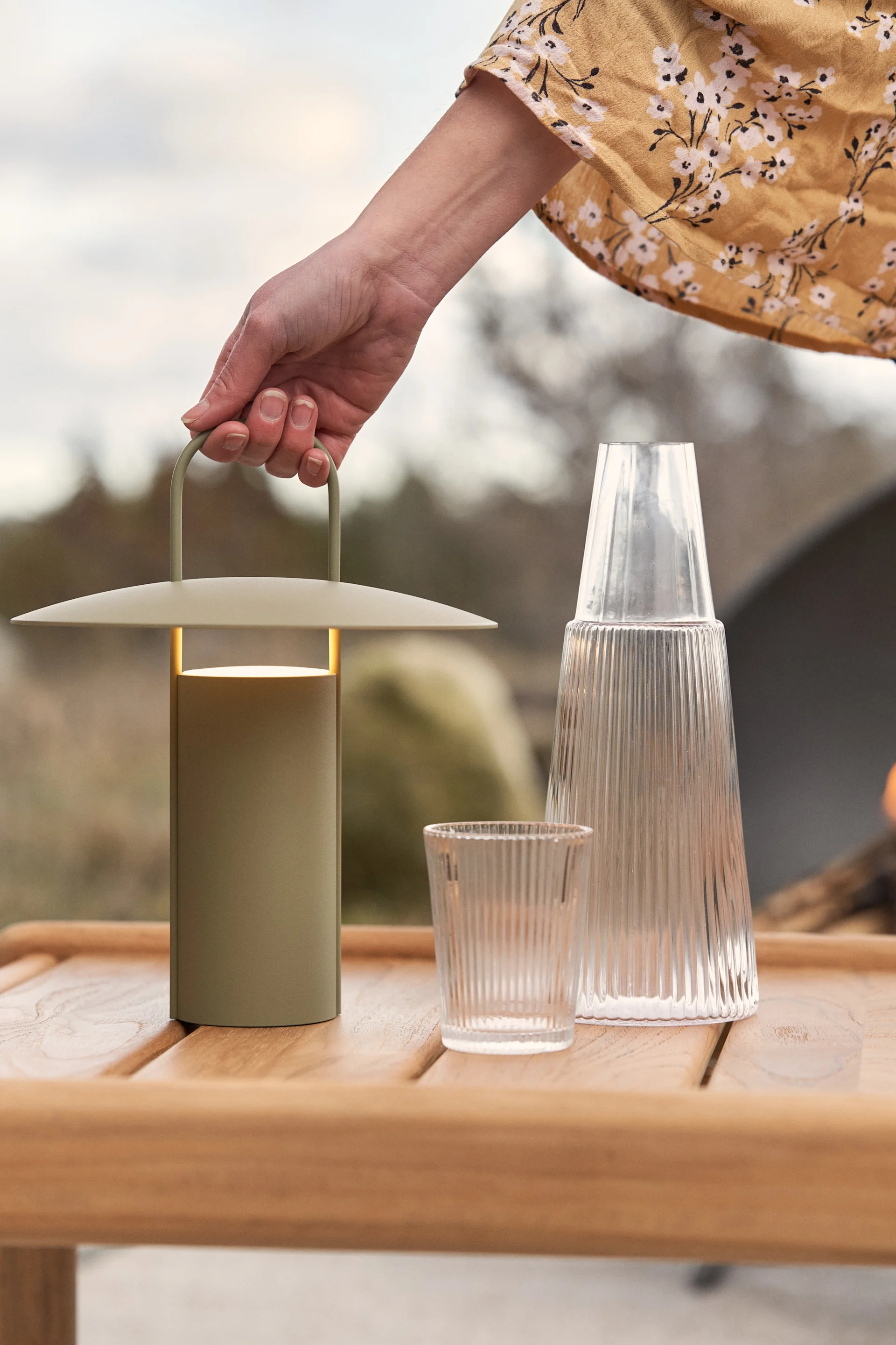 Die grüne Ray Tischleuchte von Audo Copenhagen steht draußen auf einem Holztisch, daneben eine Karaffe und ein Glas.