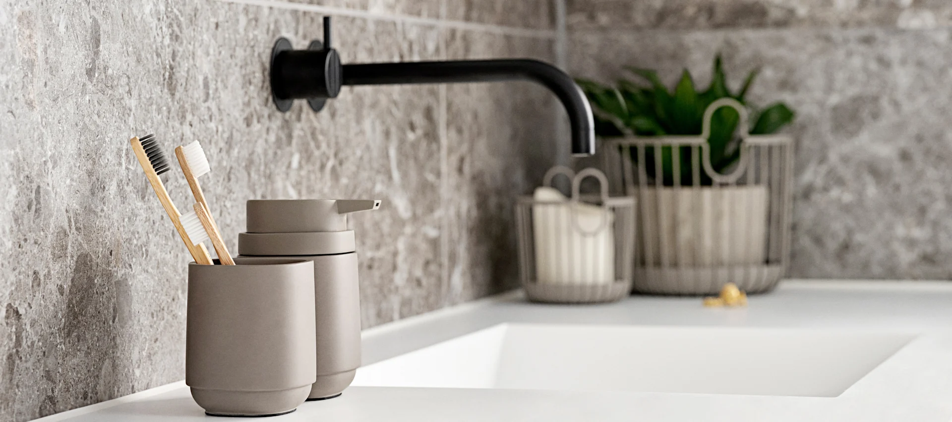 Badezimmerwaschtisch mit zwei Bambuszahnbürsten in grauem Becher, Seifenspender und schwarzem Wasserhahn.
