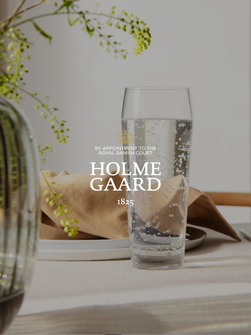 Ein hohes Glas Sprudelwasser neben einer braunen Serviette und einer Pflanze, mit dem Holmegaard-Logo überlagert.