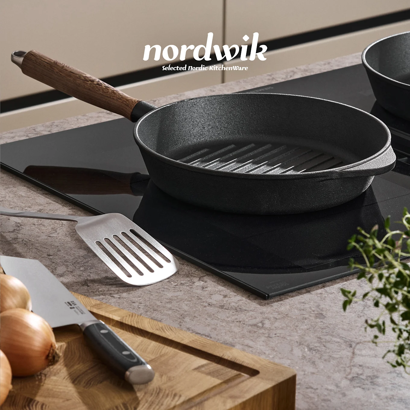 Eine schwarze Gusseisen-Grillpfanne auf einem Induktionskochfeld, mit einem Spatel, Zwiebeln, Messer und Thymian auf einem Holzbrett. Nordwik-Logo sichtbar.