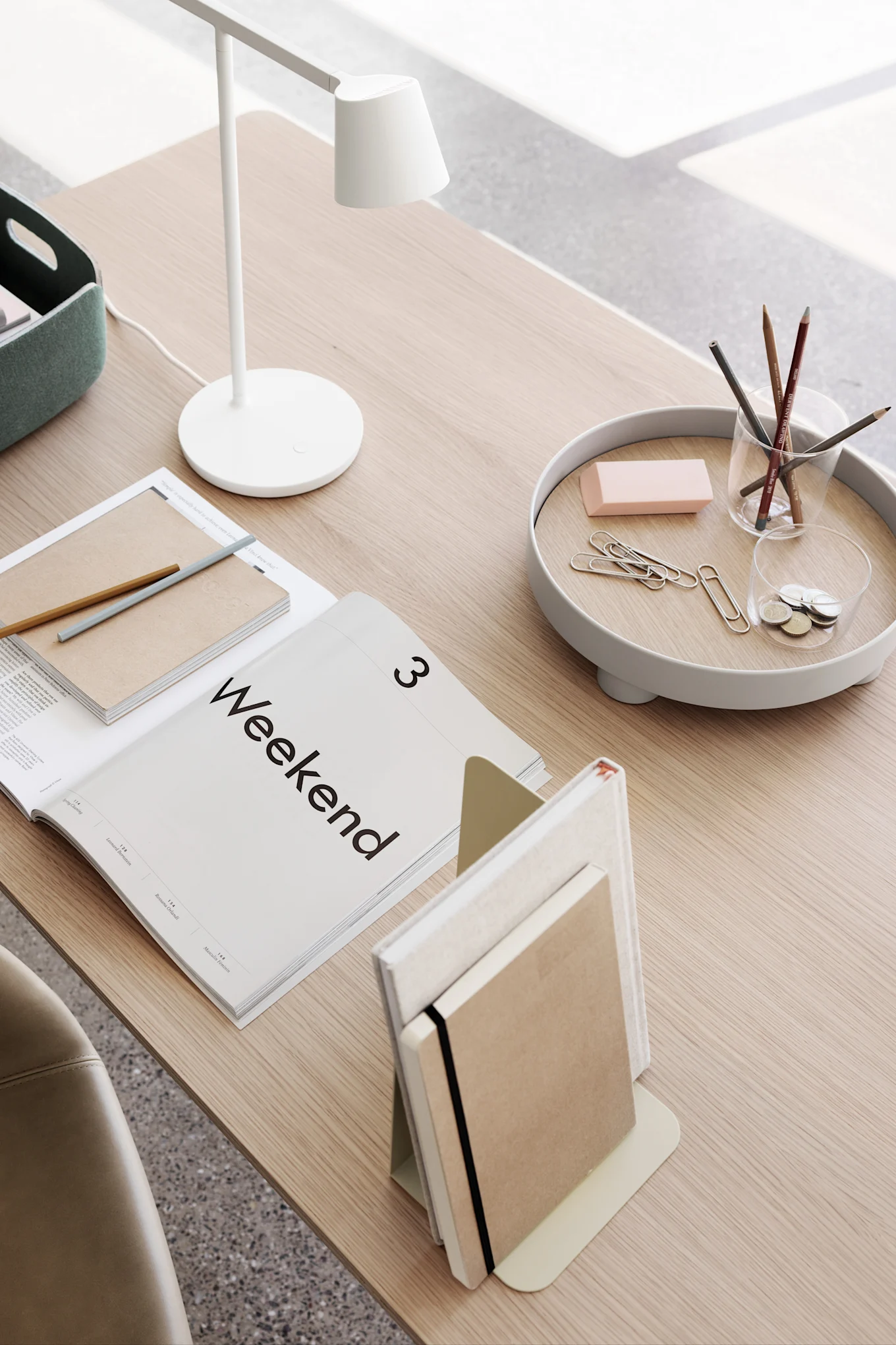 Minimalistische Schreibtischordnung mit weißer Lampe, offenem 'Weekend'-Magazin, Notizbüchern und Schreibtischzubehör auf einem Tablett.