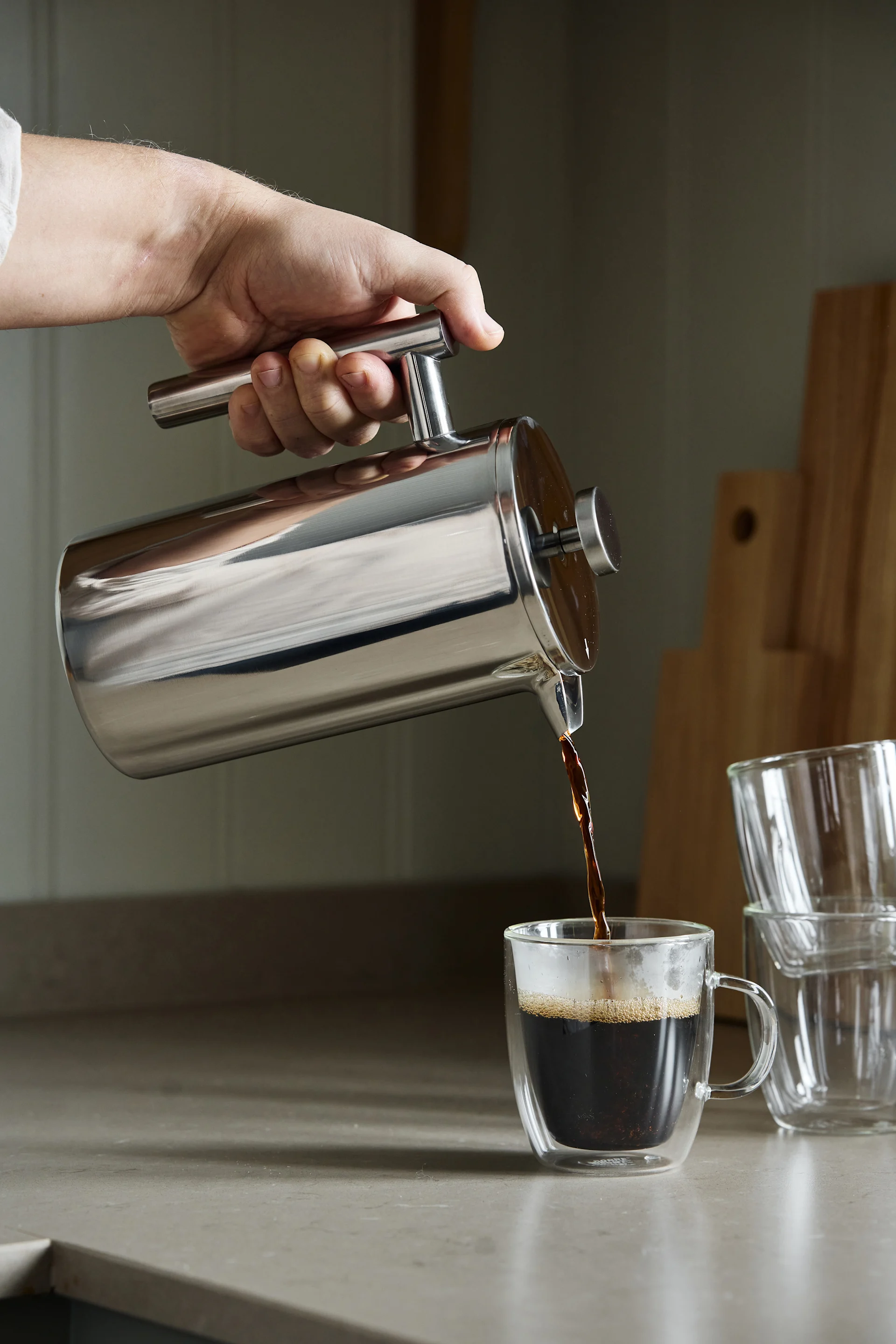 Hand gießt dunklen Kaffee aus einer glänzenden silbernen French Press in eine doppelwandige, klare Glastasse auf einer Küchentheke.