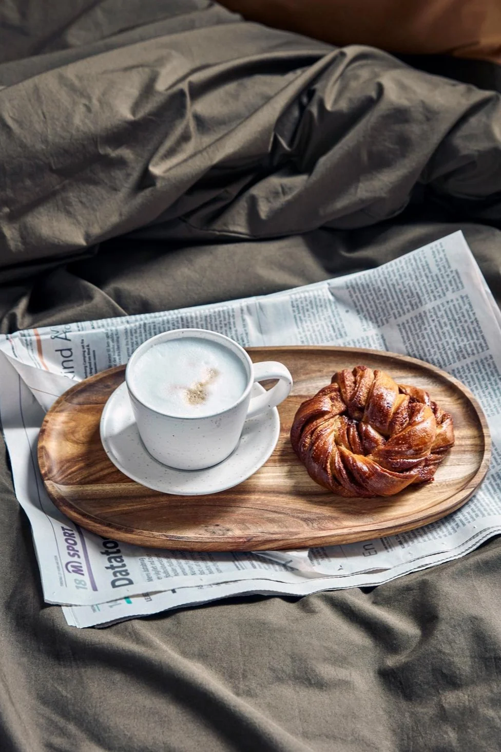 Ein Cappuccino in der weiß-grauen Pion-Tasse von House Doctor steht auf einem kleinen Holztablett, auf einer Zeitung, auf einem Bett, daneben eine schwedische Zimtschnecke.