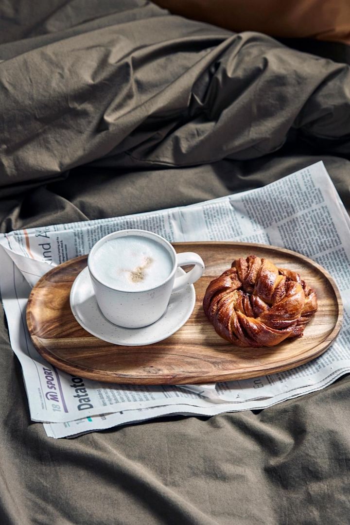 Ein Cappuccino in der weiß-grauen Pion-Tasse von House Doctor steht auf einem kleinen Holztablett, auf einer Zeitung, auf einem Bett, daneben eine schwedische Zimtschnecke.