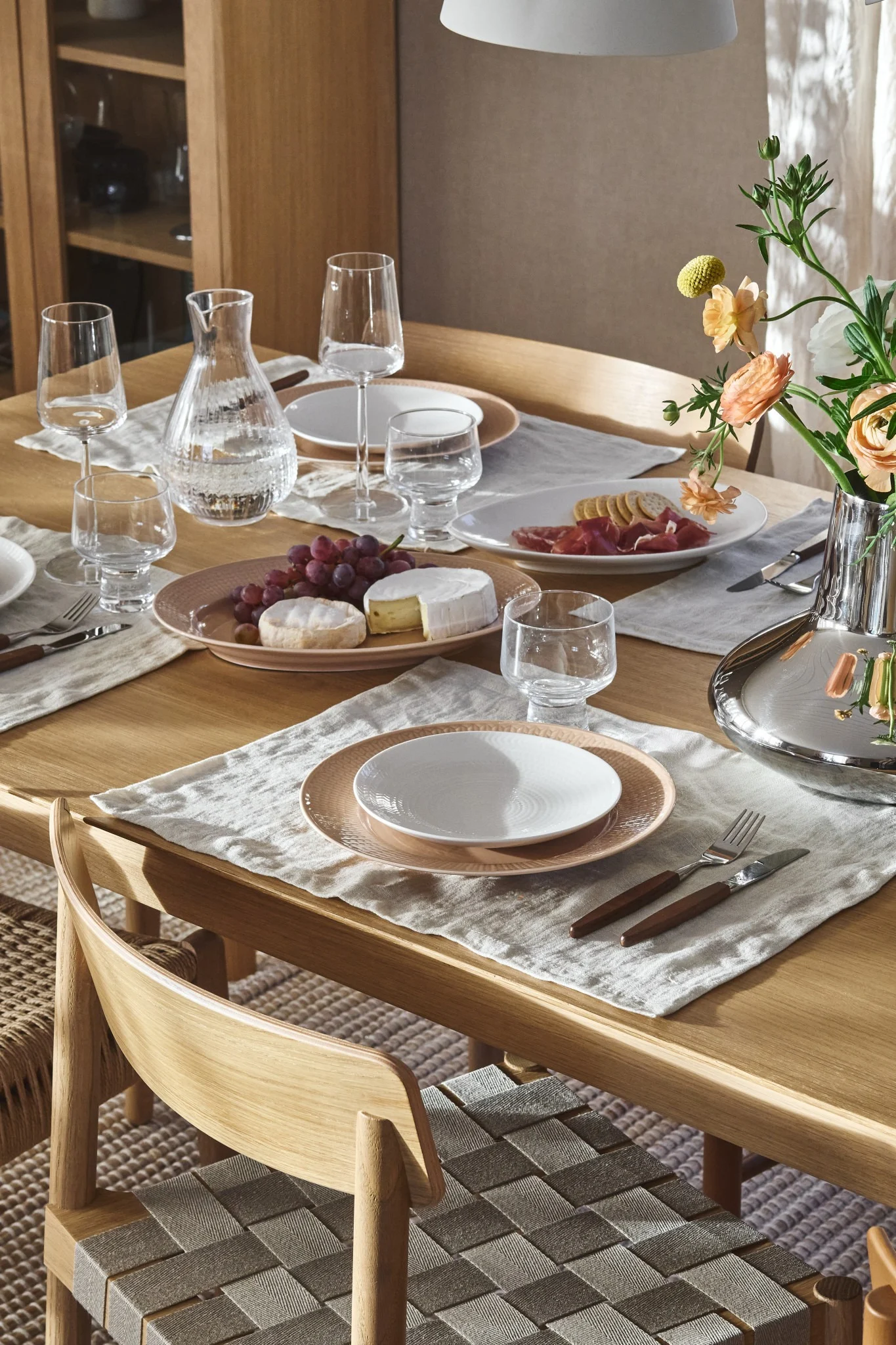 Ein Holzesstisch mit Tellern, Gläsern, Wasserkaraffe, Trauben, Käse, Wurstwaren und Blumen. Geflochtene Stühle.