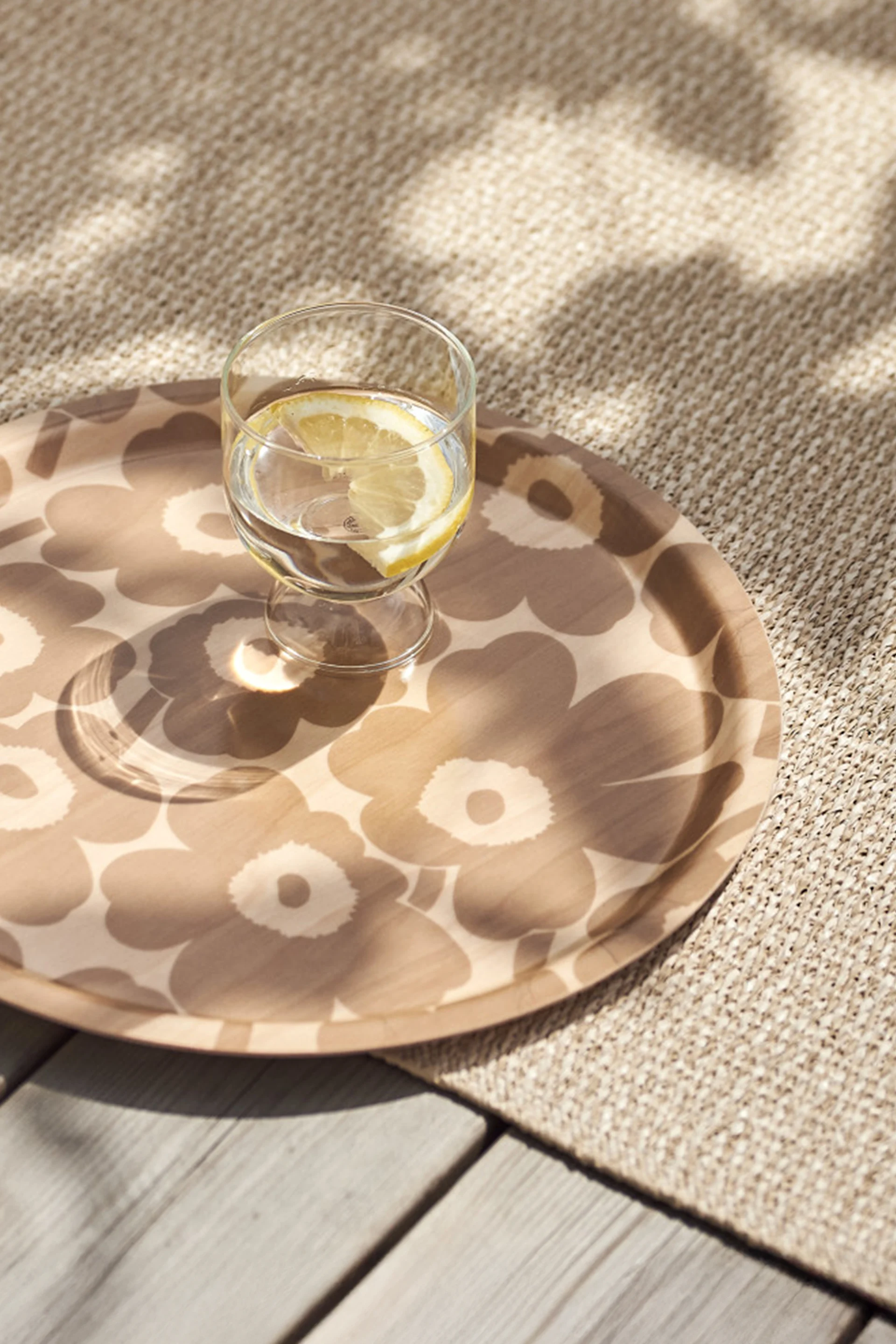 Ein Glas Wasser mit Zitronenscheibe auf einem gemusterten runden Tablett, platziert auf einem beigefarbenen Teppich und Holzdeck mit geflecktem Sonnenlicht.