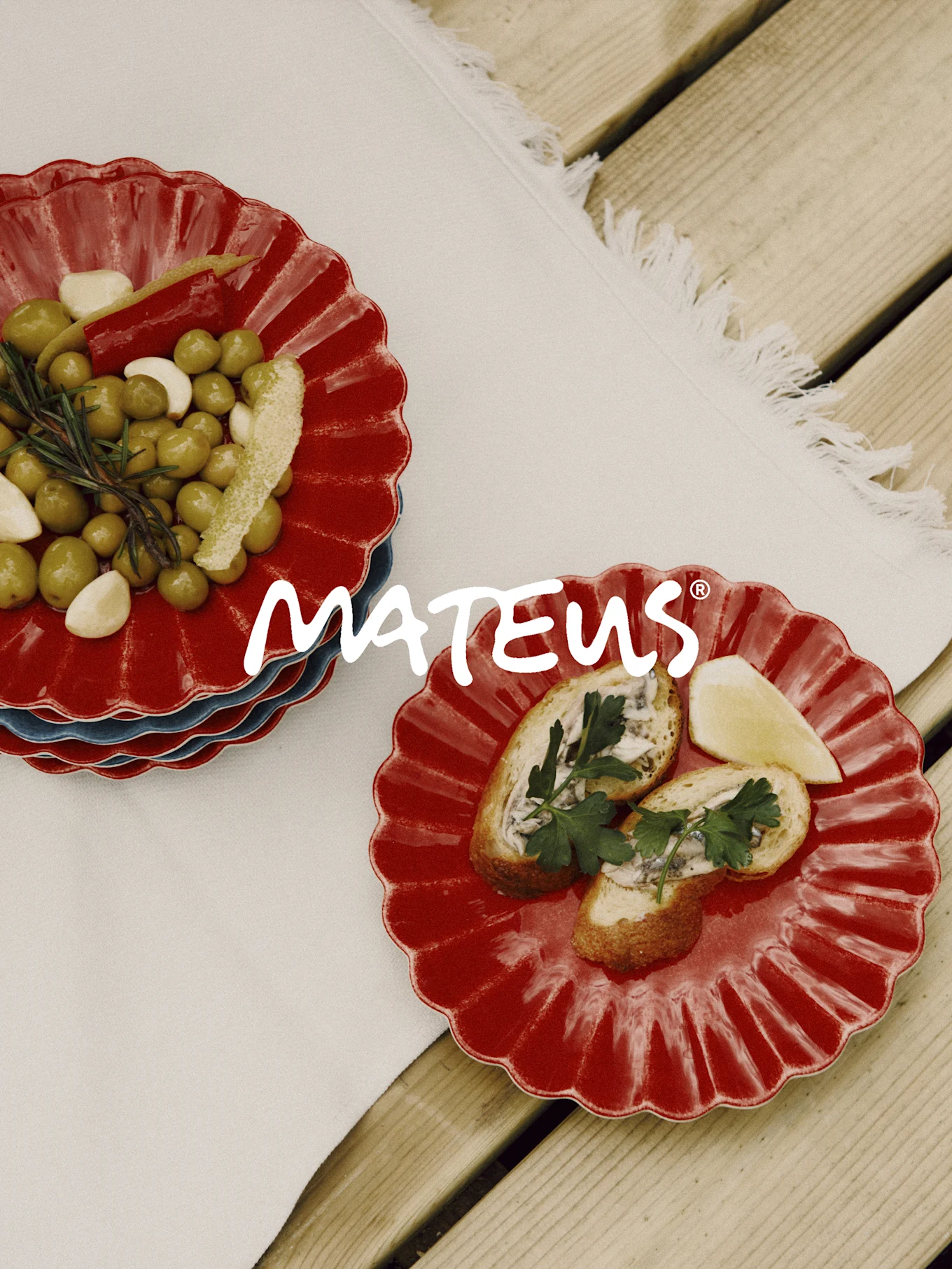 Rote Teller mit Jakobsmuschelrand, darauf Oliven, Knoblauch, Rosmarin und Brot mit Sardellen. Marke MATEUS.