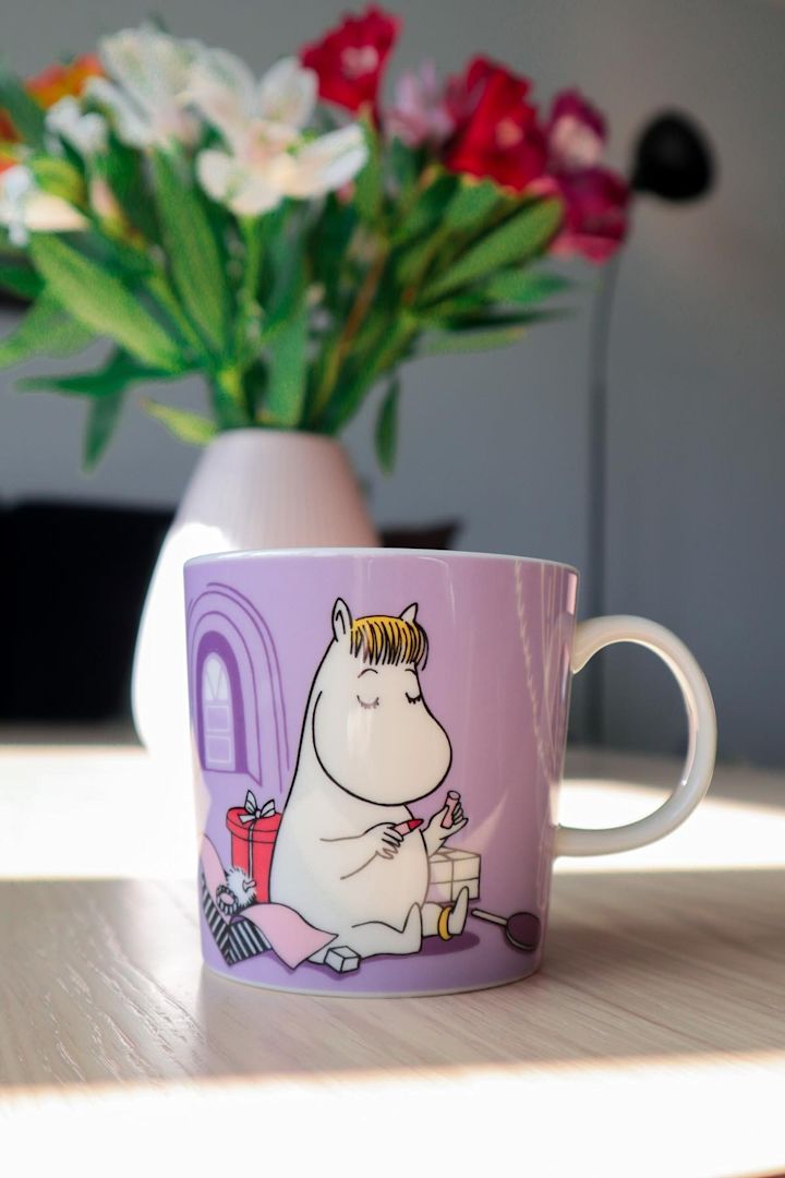 Die lilafarbene Mumin-Tasse mit dem Snorkfräulein darauf steht auf einem Tisch, im Hintergrund steht eine Vase mit einem Strauß Blumen.