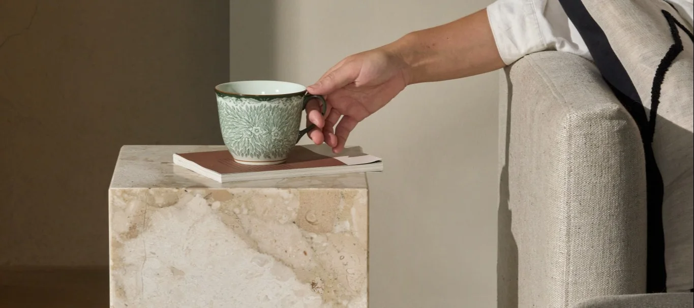 Eine Hand nimmt eine grüne, gemusterte Tasse von einem Stapel Notizbücher auf einem Marmor-Beistelltisch neben einem Sessel.