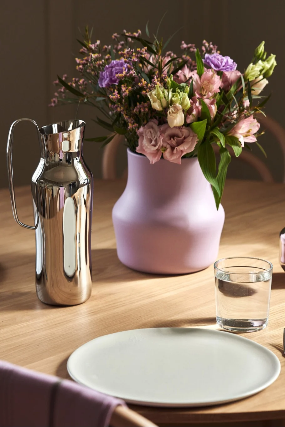 Hier sehen Sie die violette Dorotea-Vase mit einem pink- und lilafarbenen Blumenstrauß neben der silbernen Dorotea Karaffe, beides von Gense, auf einem Tisch stehen.