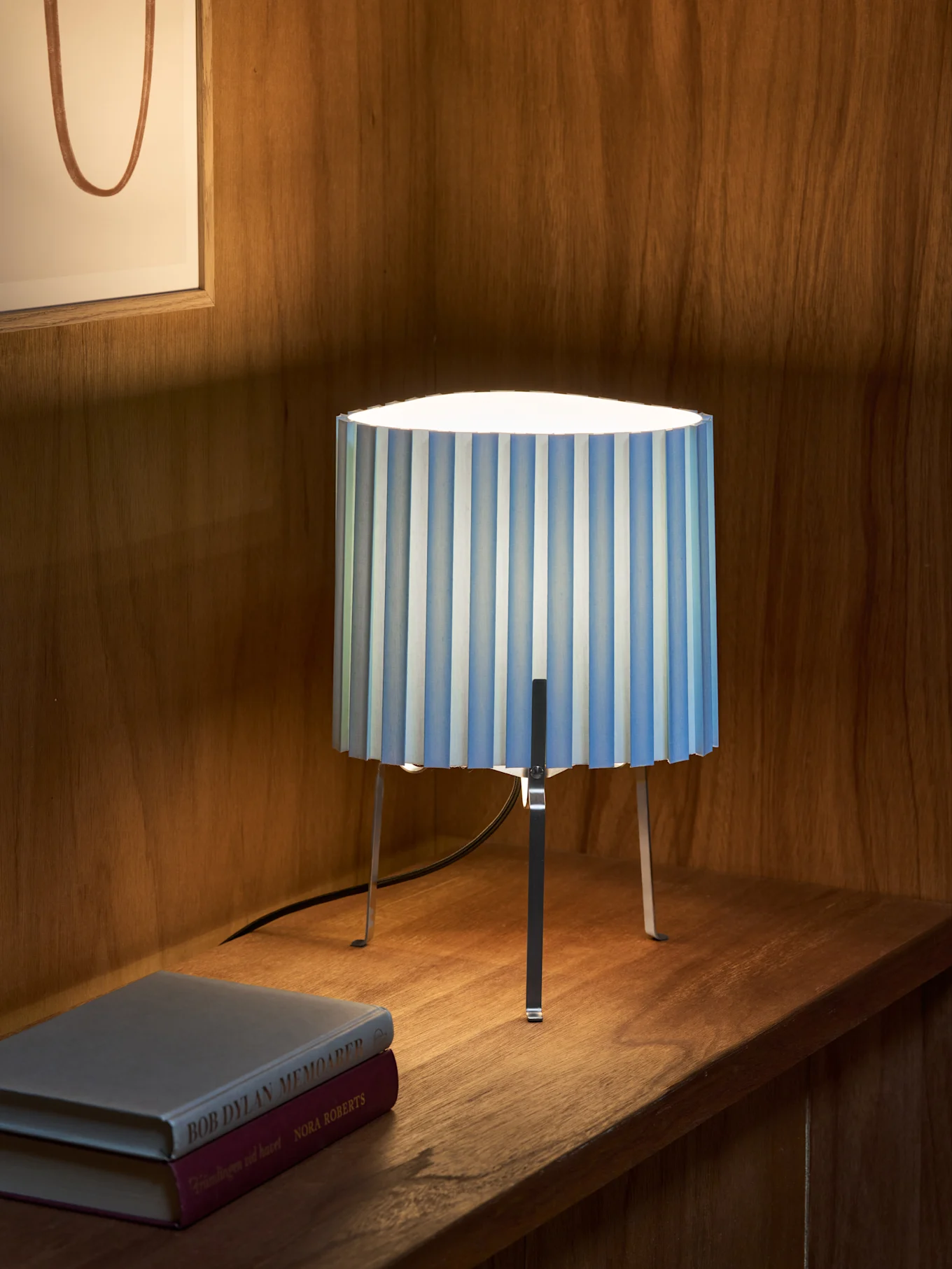 Eine blau-weiß gestreifte, plissierte Tischlampe mit drei Metallbeinen steht auf einem Holzregal neben zwei Büchern. Gerahmtes Kunstwerk im Hintergrund.