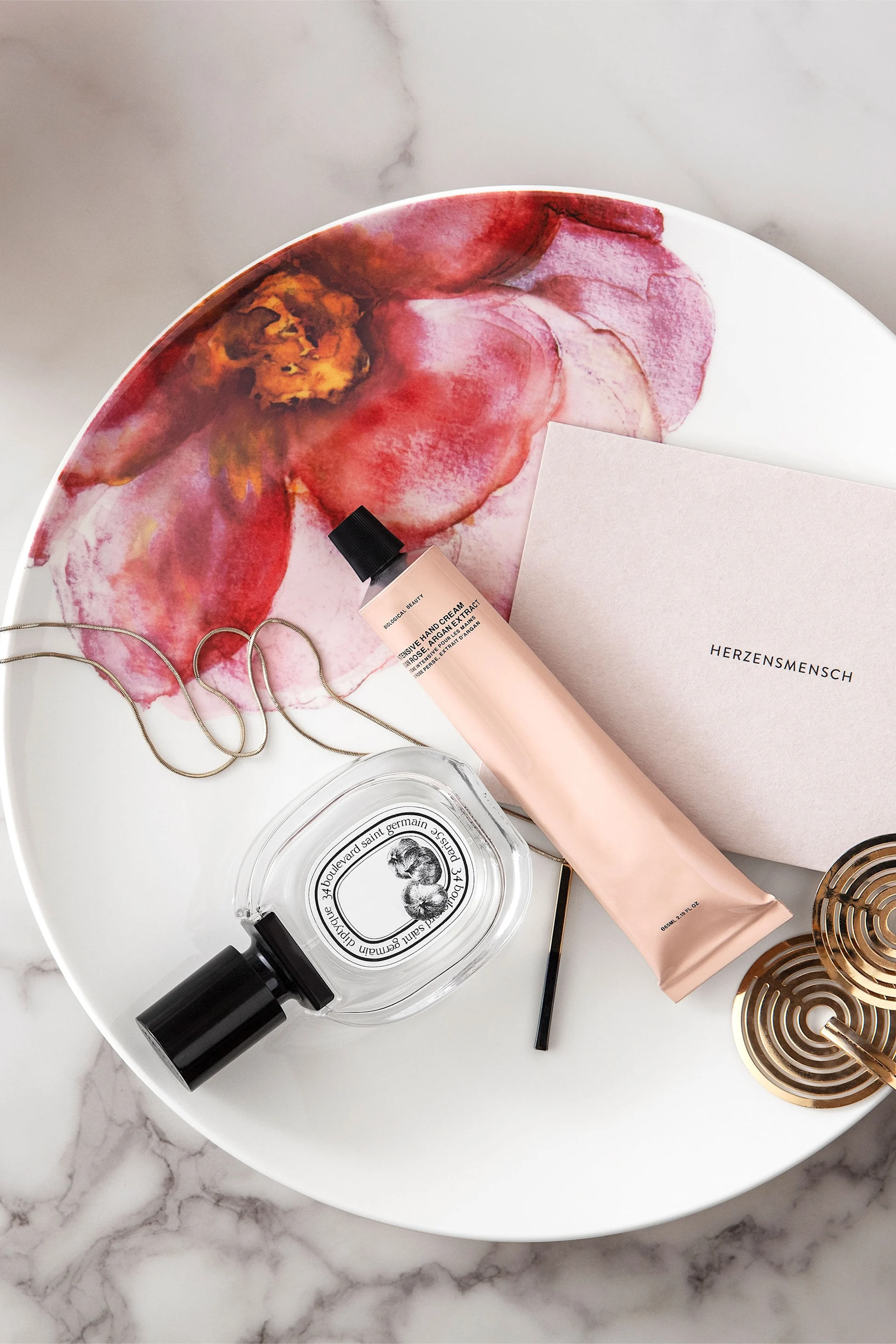 Parfüm, pfirsichfarbene Handcreme, Goldkette und Untersetzer auf einem weißen Tablett mit pinkem Blumendesign.