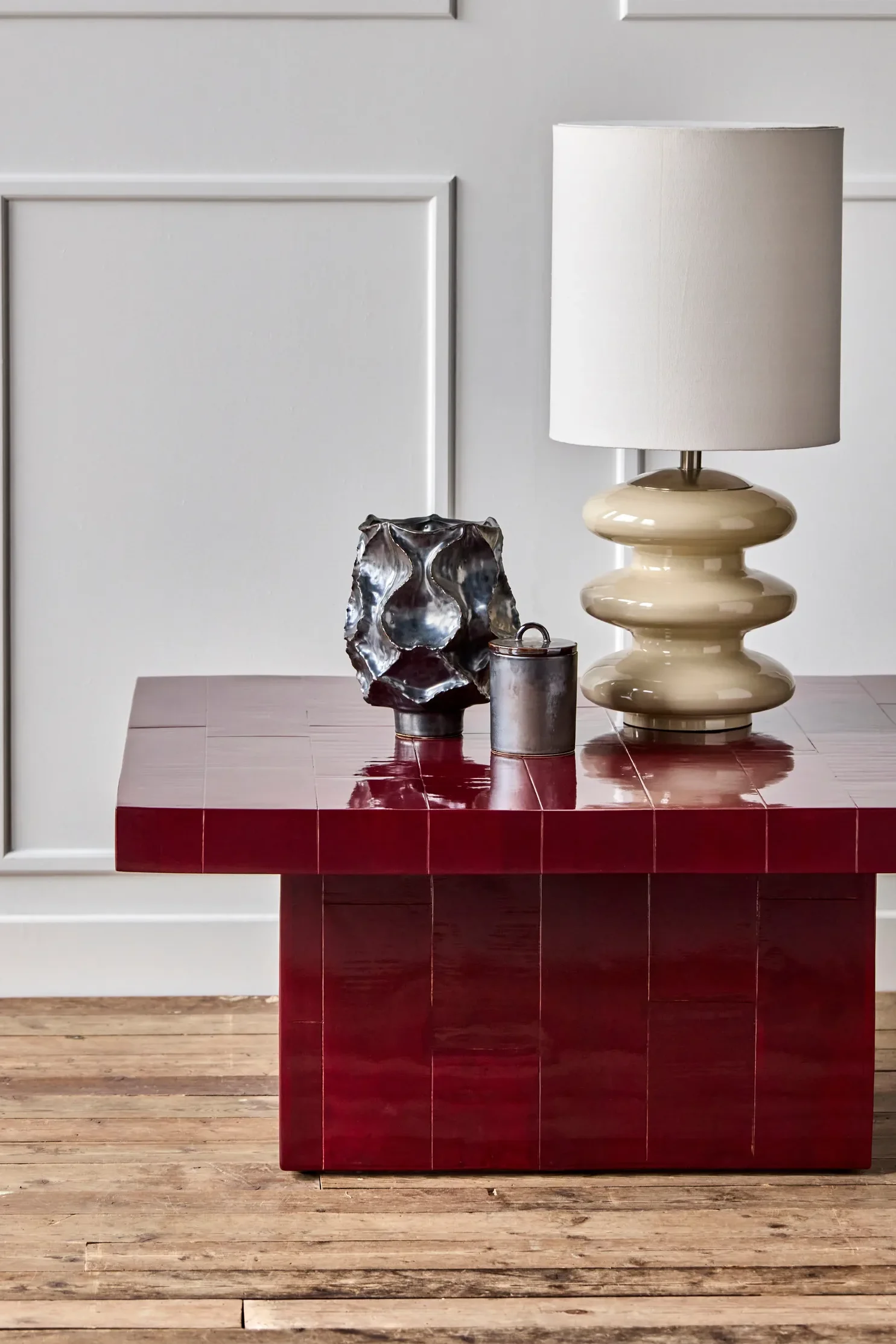 Glänzender dunkelroter Fliesencouchtisch mit cremefarbener Skulpturlampe, dunkler metallischer abstrakter Vase und kleinem zylindrischem Behälter.