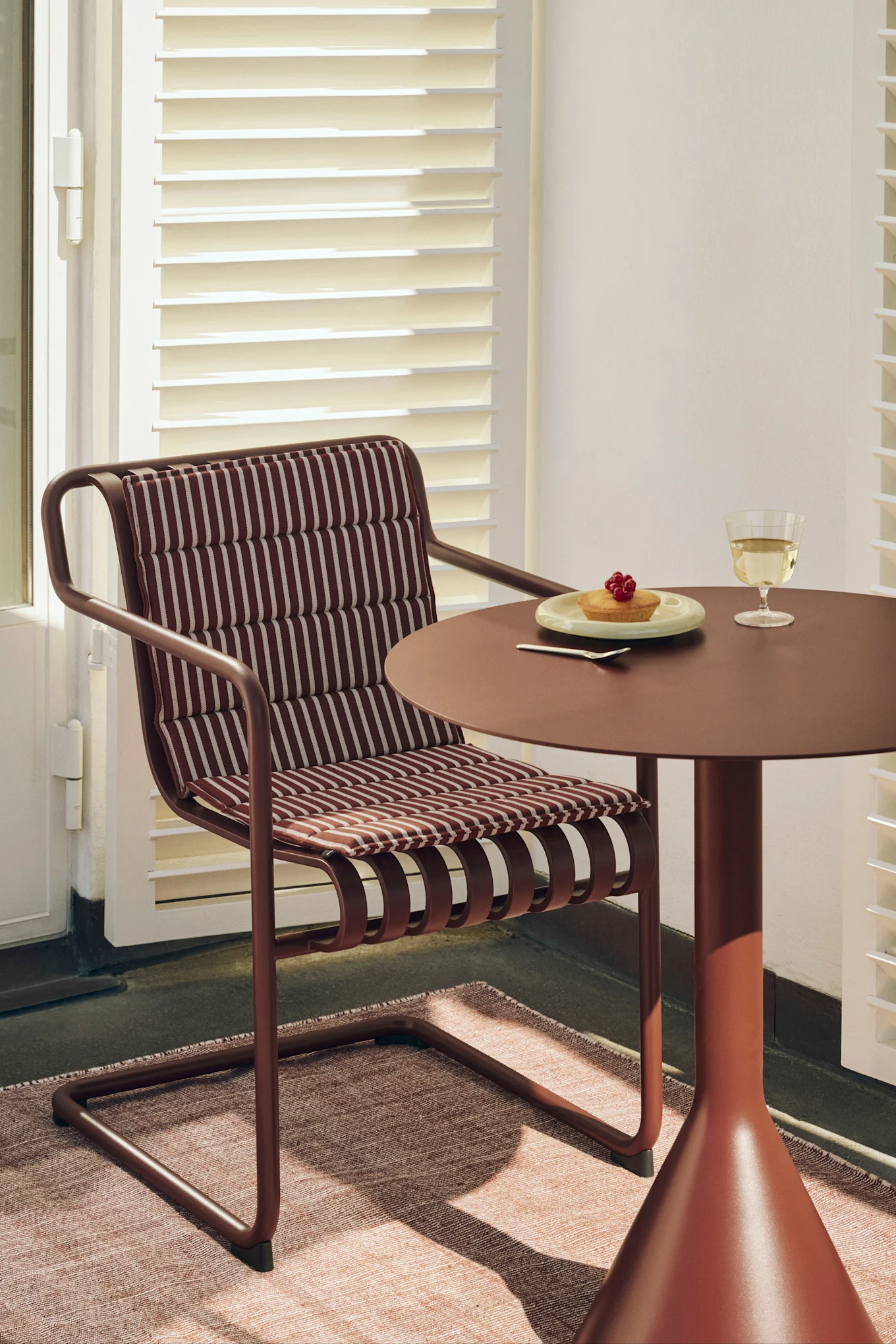 Ein rotbrauner Stuhl mit gestreiftem Kissen an einem passenden runden Tisch mit Kuchen und Weißwein.