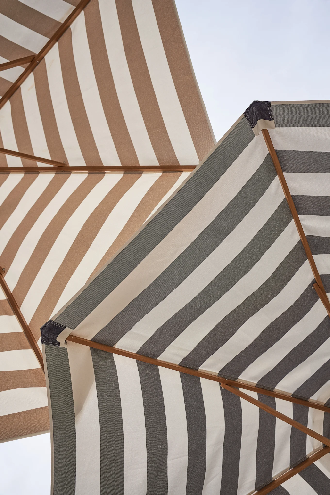 Zwei gestreifte Sonnenschirme von unten gesehen, einer braun-weiß, der andere grau-weiß, vor hellem Himmel.