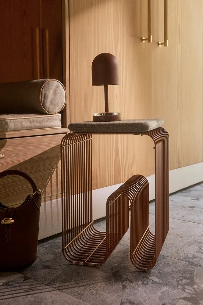 Moderner Beistelltisch aus braunen vertikalen Metallstangen mit brauner Kuppellampe und Handtasche, vor einem Holzschrank.