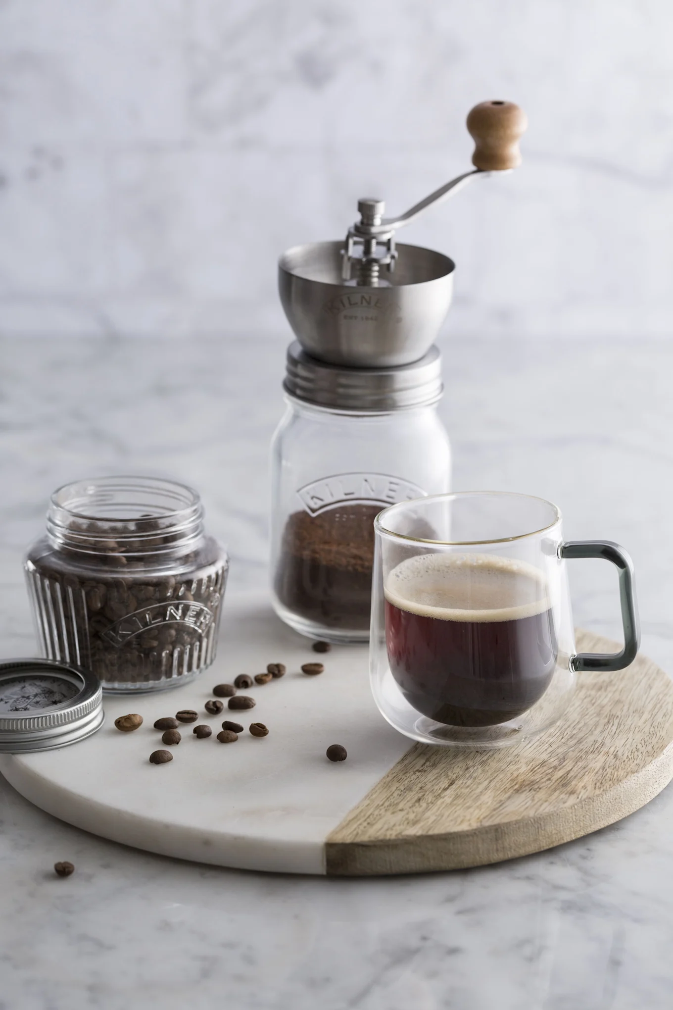 Manuelle Kaffeemühle über gemahlenem Kaffee, ganze Bohnen im Glas und eine Tasse schwarzen Kaffee auf einer zweifarbigen Marmor- und Holzplatte.