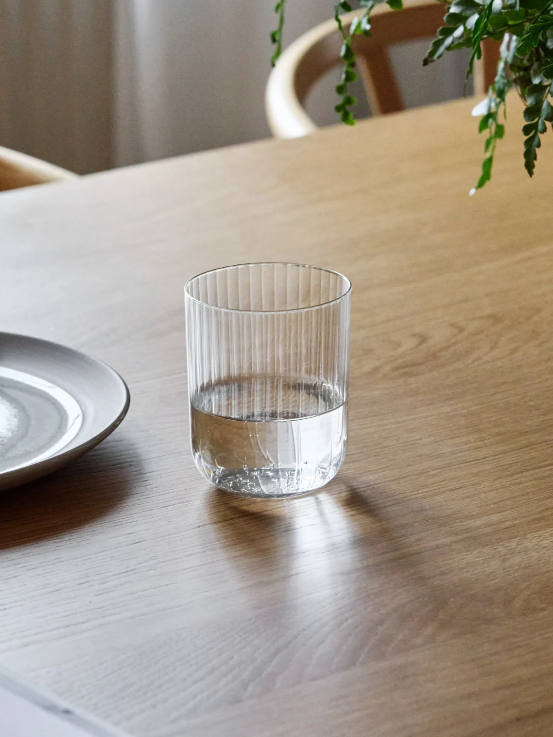 Ein geripptes Glas, halb mit Wasser gefüllt, steht auf einem Holztisch neben einem Keramikteller.