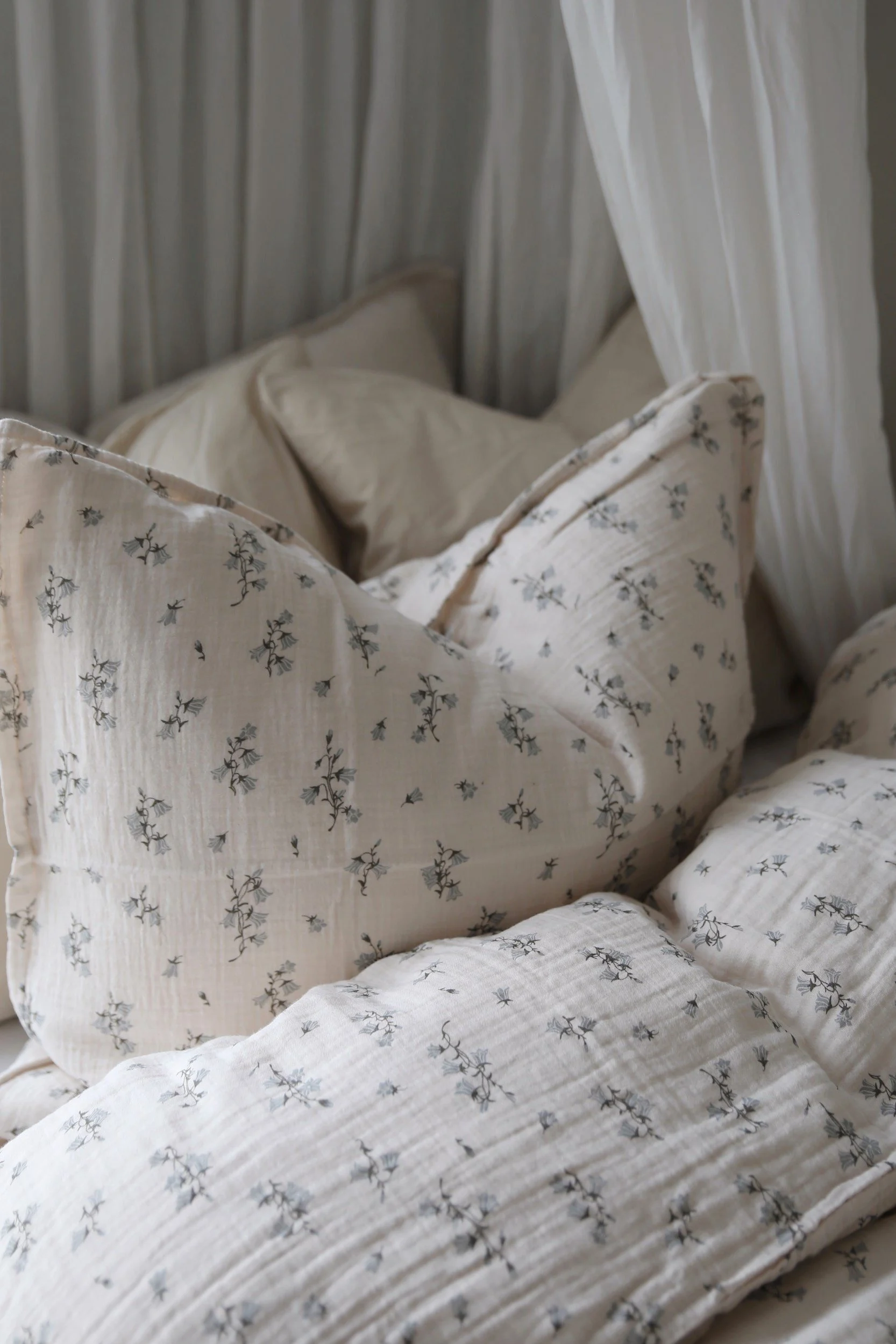 Gemütliche Bettwäsche aus hellem, beigefarbenem Crinkle-Stoff mit zartem blauem Blumenmuster und weißen Vorhängen.