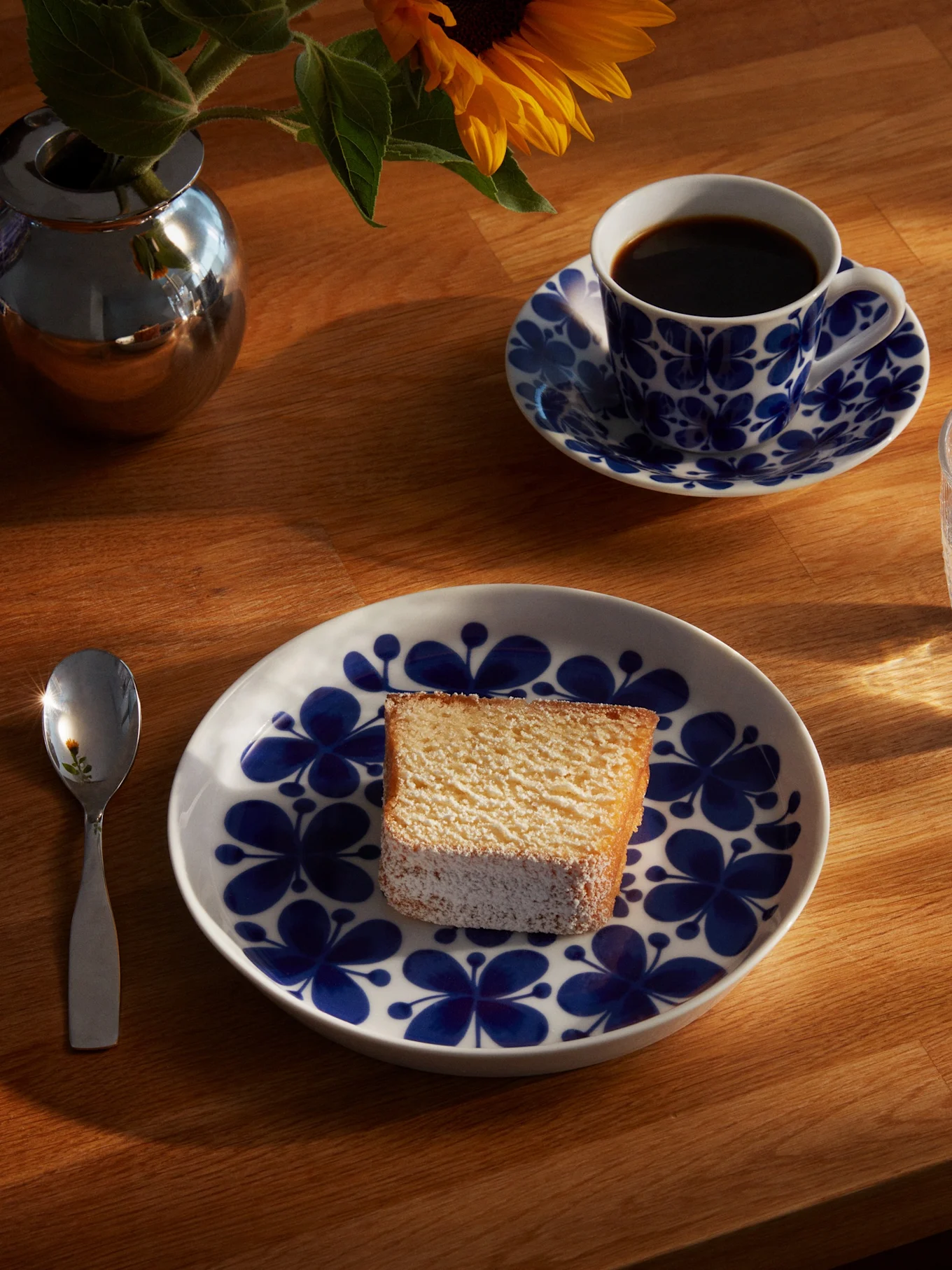 Ein Stück Kuchen auf einem blau-weiß gemusterten Teller, dazu eine Kaffeetasse, ein Glas Wasser, ein Löffel und eine Sonnenblume in einer silbernen Vase auf einem sonnenbeschienenen Holztisch.