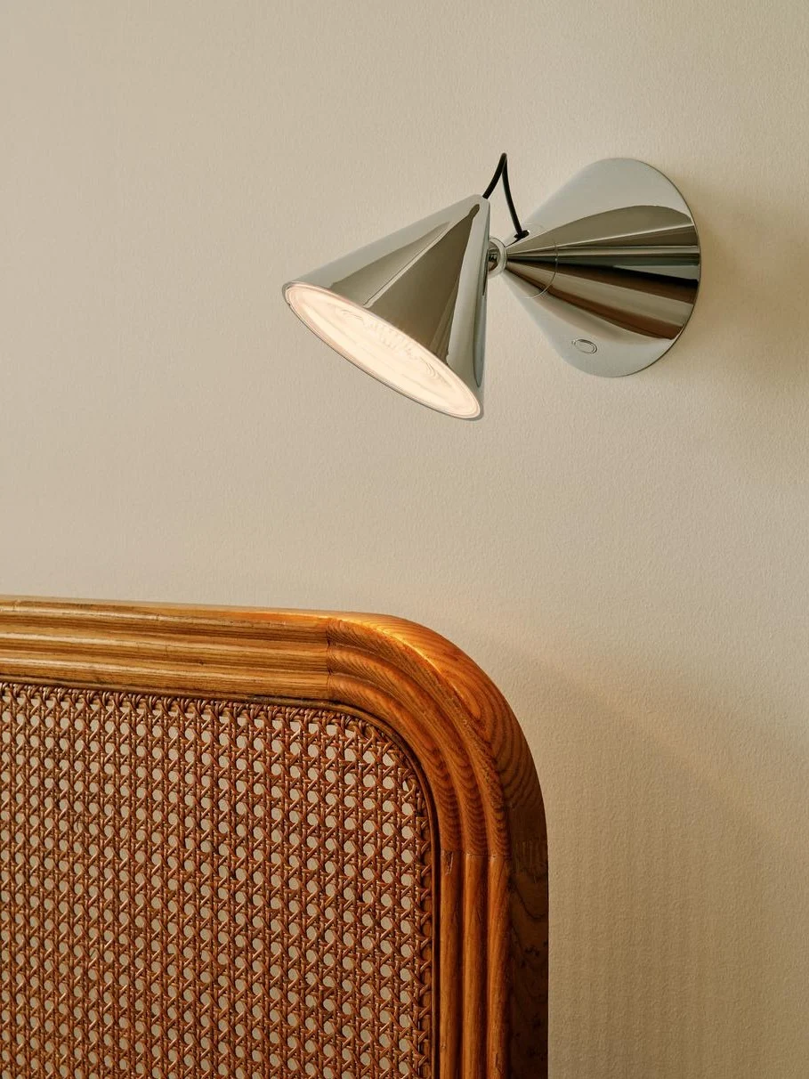Moderne Chrom-Wandleuchte mit konischem Schirm, die eine helle Wand über einem geflochtenen Rattan- und Holzkopfteil beleuchtet.