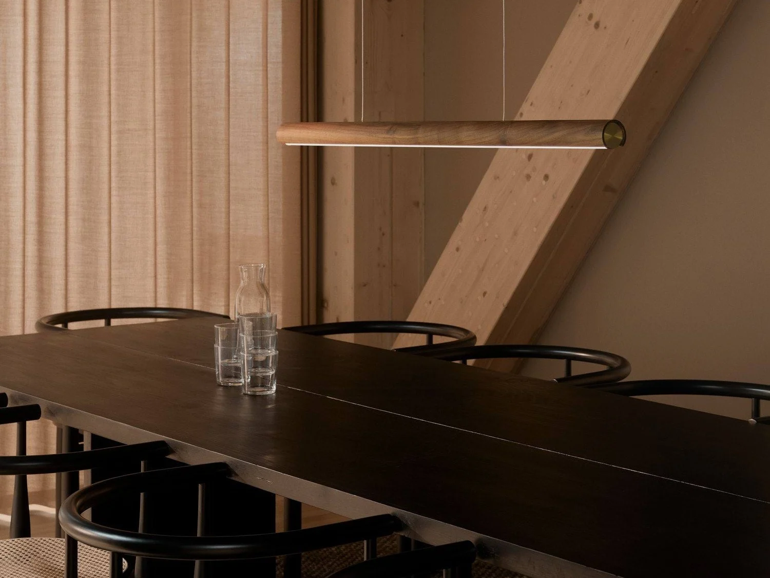 Ein modernes Esszimmer mit einem langen dunklen Holztisch, schwarzen Stühlen und einer schlanken Pendelleuchte aus Holz. Eine Glaskaraffe und drei Gläser stehen auf dem Tisch.