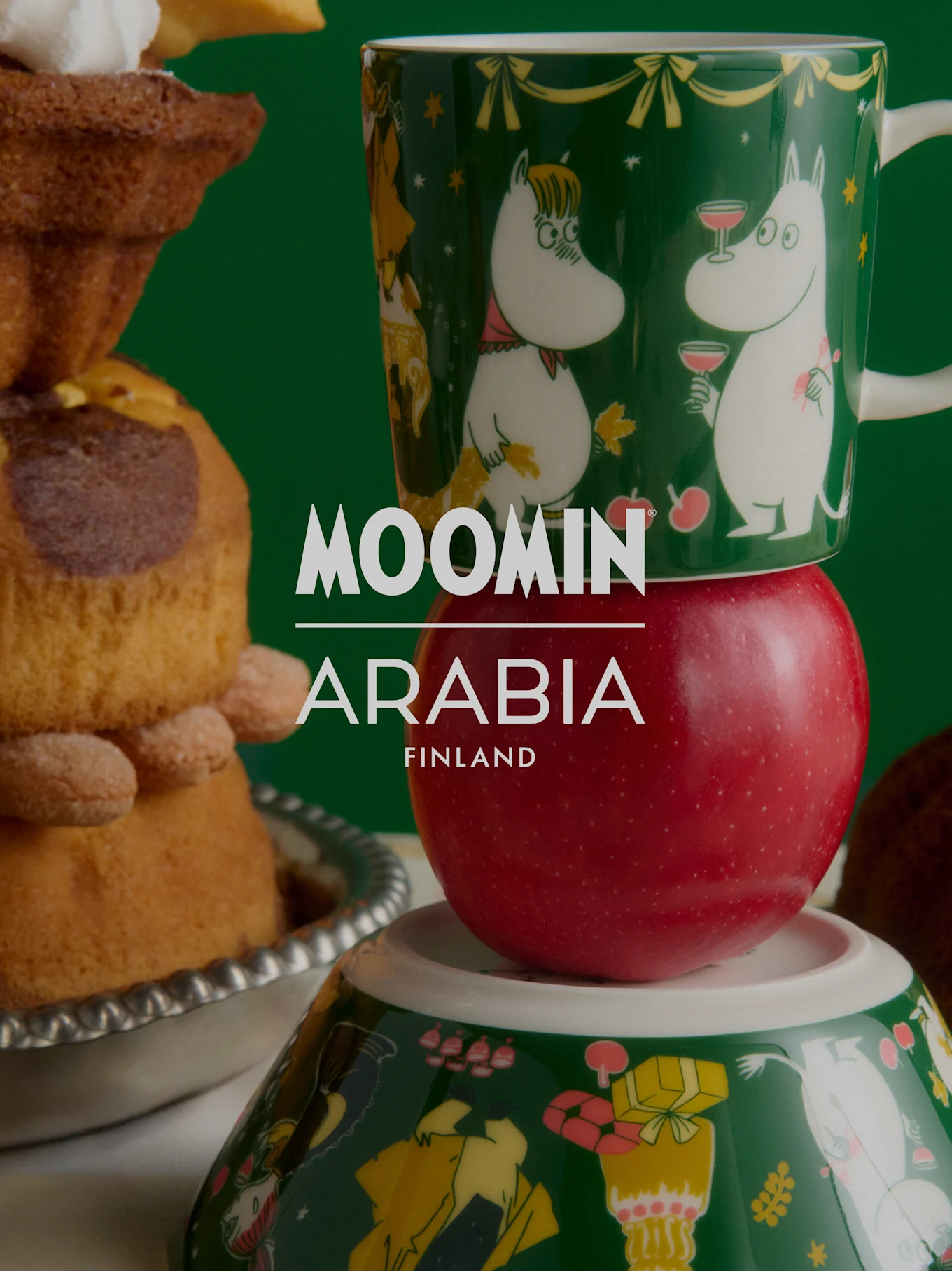 Moomin Arabia Weihnachtskollektion: Eine grüne Moomin-Tasse auf einem roten Apfel, über einer Moomin-Schale, neben festlichem Gebäck.