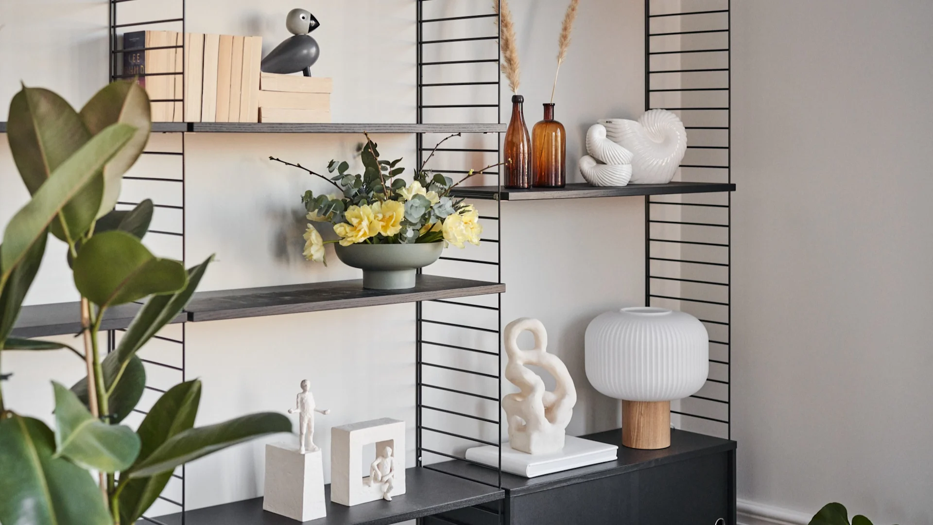Schwarzes Regal mit Büchern, Vogelfigur, gelben Blumen, Vasen, weißen Skulpturen und Lampe; grüne Pflanze vorn.