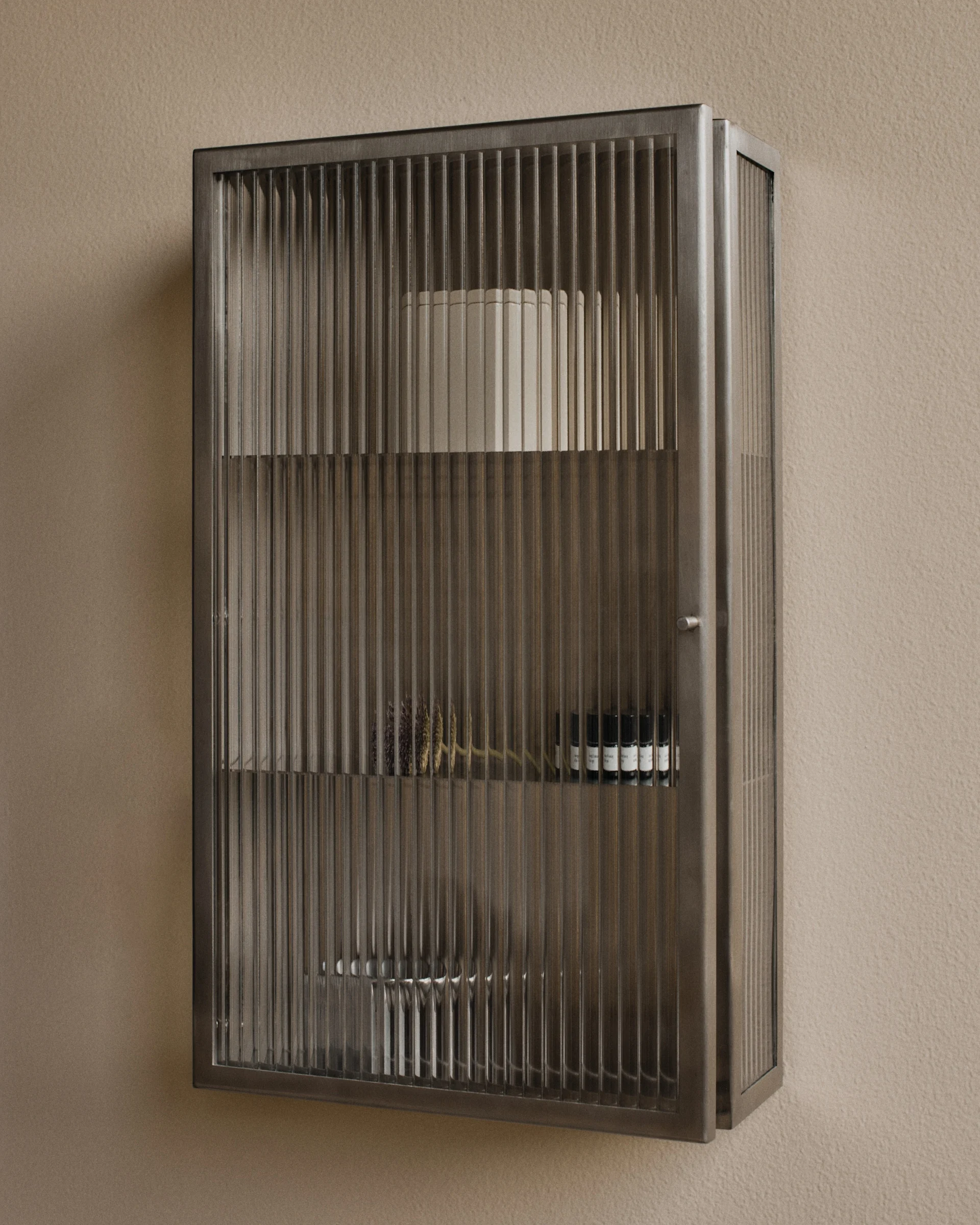 Wandmontierter Metallschrank mit geriffelten Glastüren, gefüllt mit Dekoartikeln und kleinen Flaschen.
