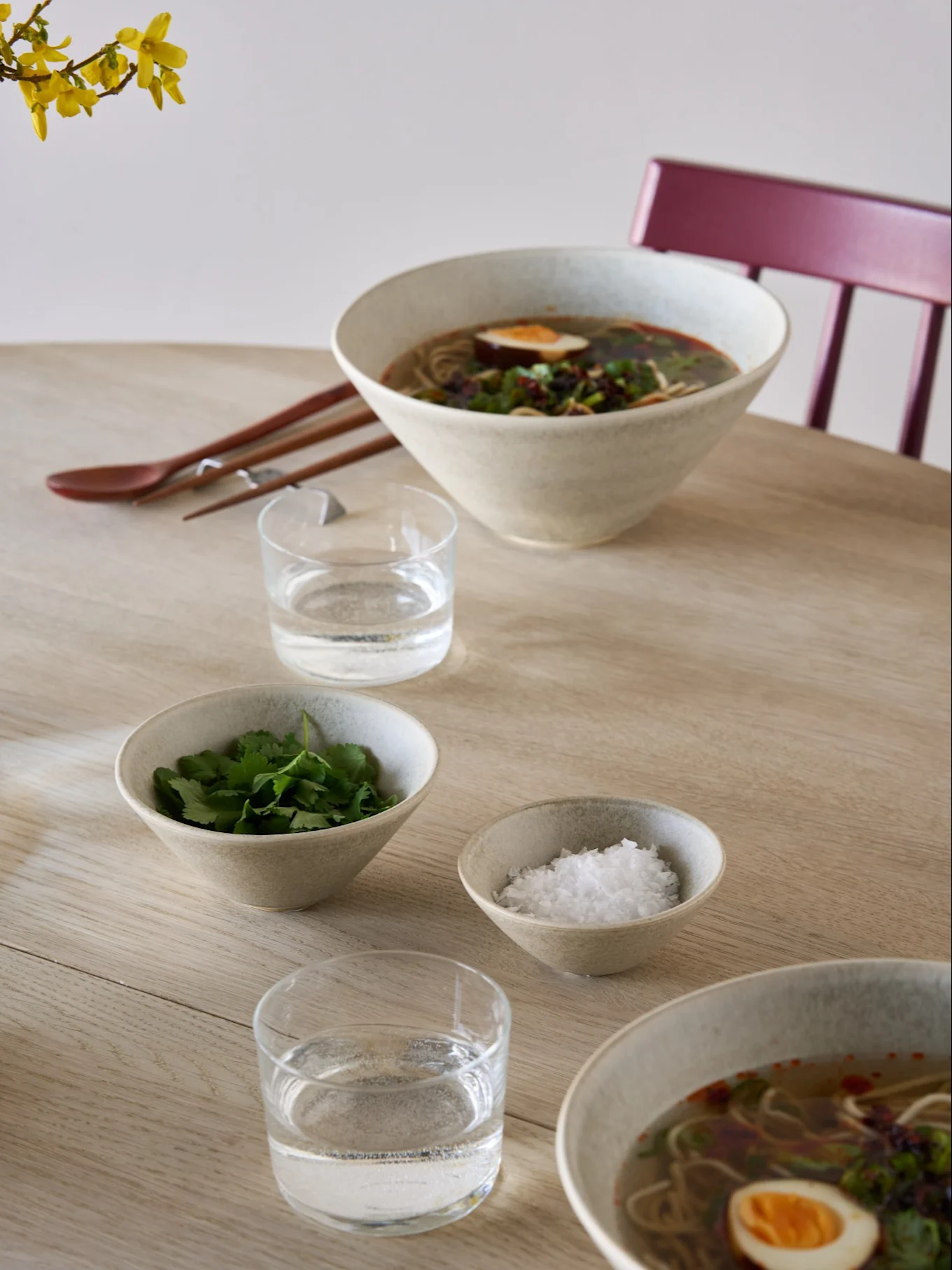 Zwei Schalen Ramen mit Ei und Kräutern, Beilagenschalen mit Koriander und Salz, Wasser, Essstäbchen und einem Löffel auf einem hellen Holztisch.