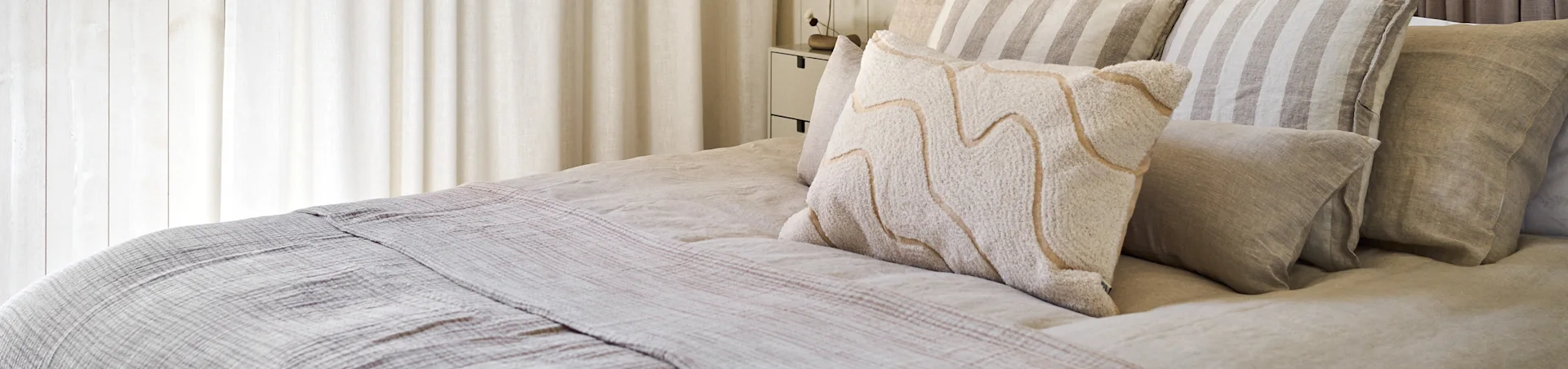 Ein Bett mit neutral getönter Leinenbettwäsche, gestreiften Kissen und einem strukturierten weißen Kissen mit wellenförmigen beigen Linien.