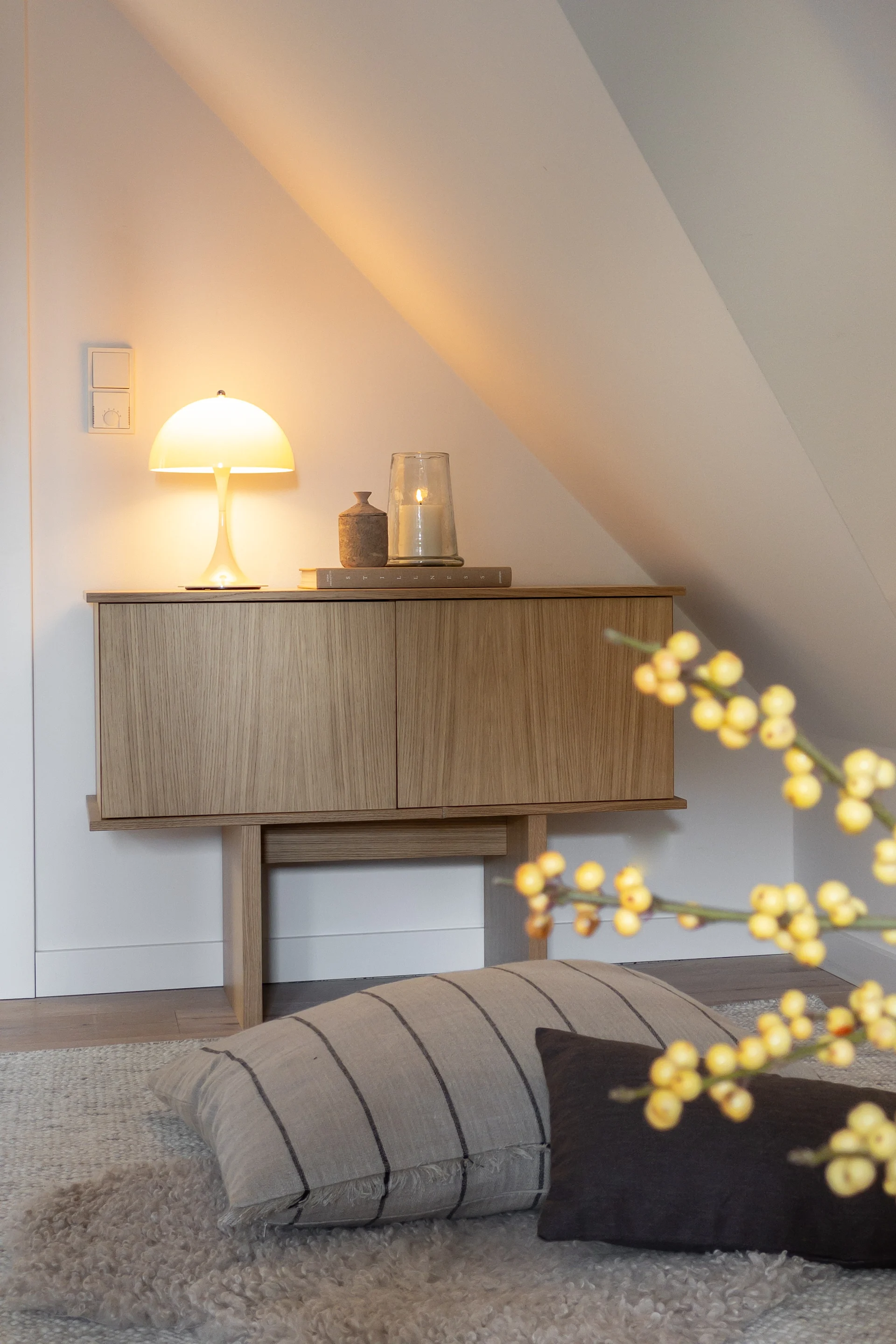 Ein Holzschrank mit einer leuchtenden Pilzlampe und einer brennenden Kerze vor einer weißen, schrägen Wand in einem gemütlichen Raum.