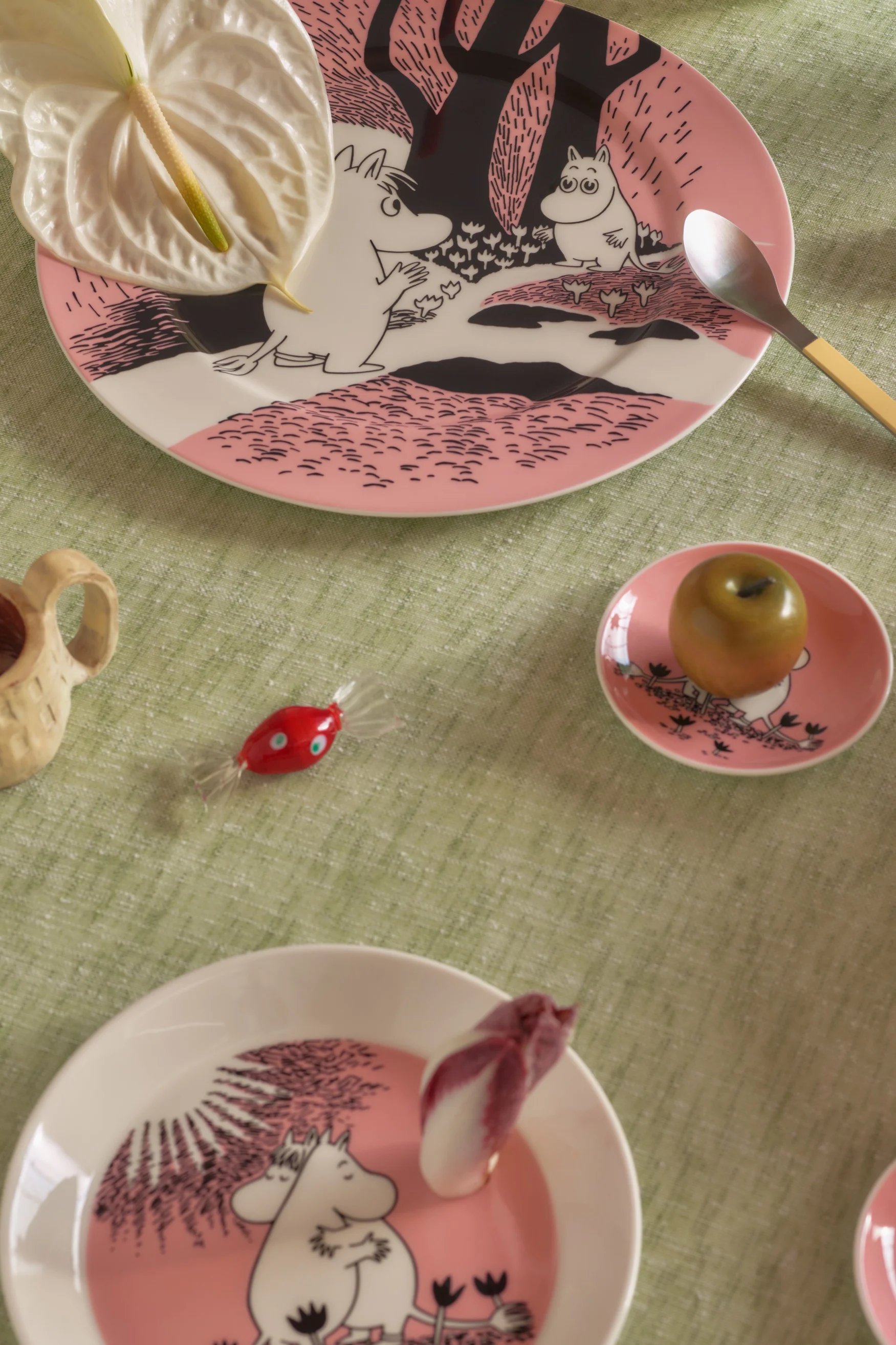 Tisch gedeckt mit rosa Mumin-Tellern, Anthurienblüte, Löffel, grünem Apfel, rotem Bonbon und einem Holzbecher auf einer grünen Tischdecke.