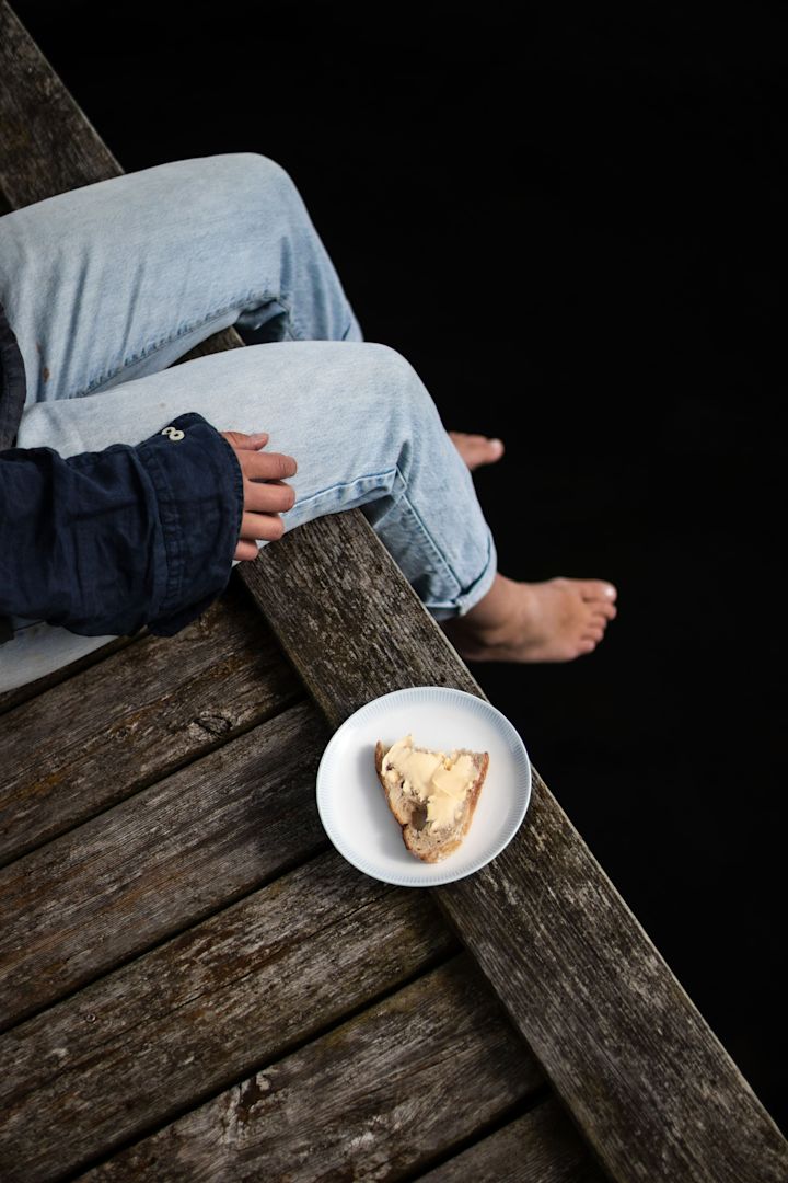 Lagom: Eine Frau sitzt auf einem Steg, daneben ein Osean Teller von Wik&Walsoe mit einem Butterbrot.