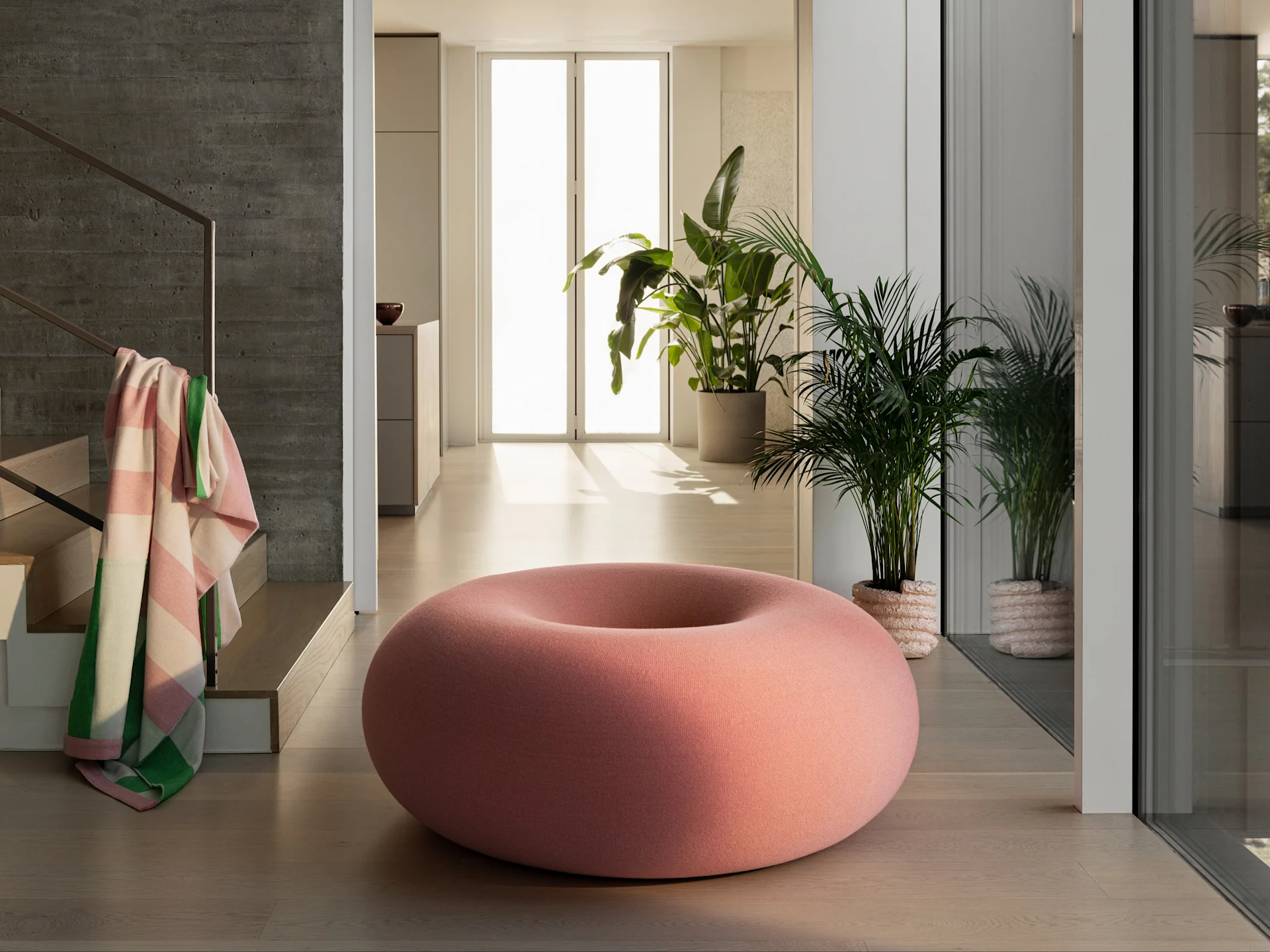 Ein großer, donut-förmiger pinker Pouf auf hellem Holzboden in einem modernen Raum mit Grünpflanzen und einer Betonwand.