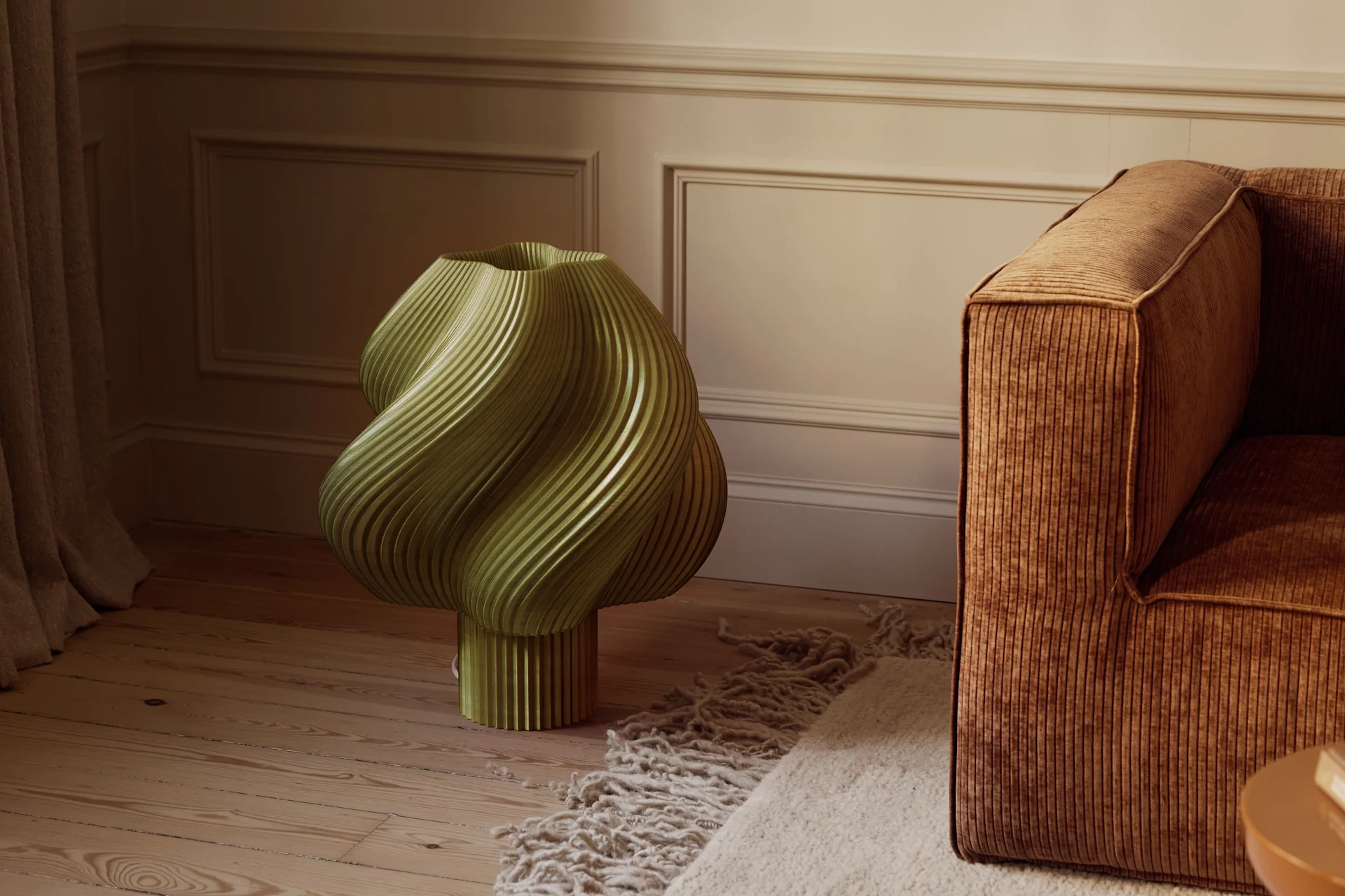 Ein olivgrünes, skulpturales, geriffeltes Objekt auf einem Holzfußboden neben einem braunen Kordsofa und einem strukturierten Teppich in einem Raum mit profilierten Wänden.