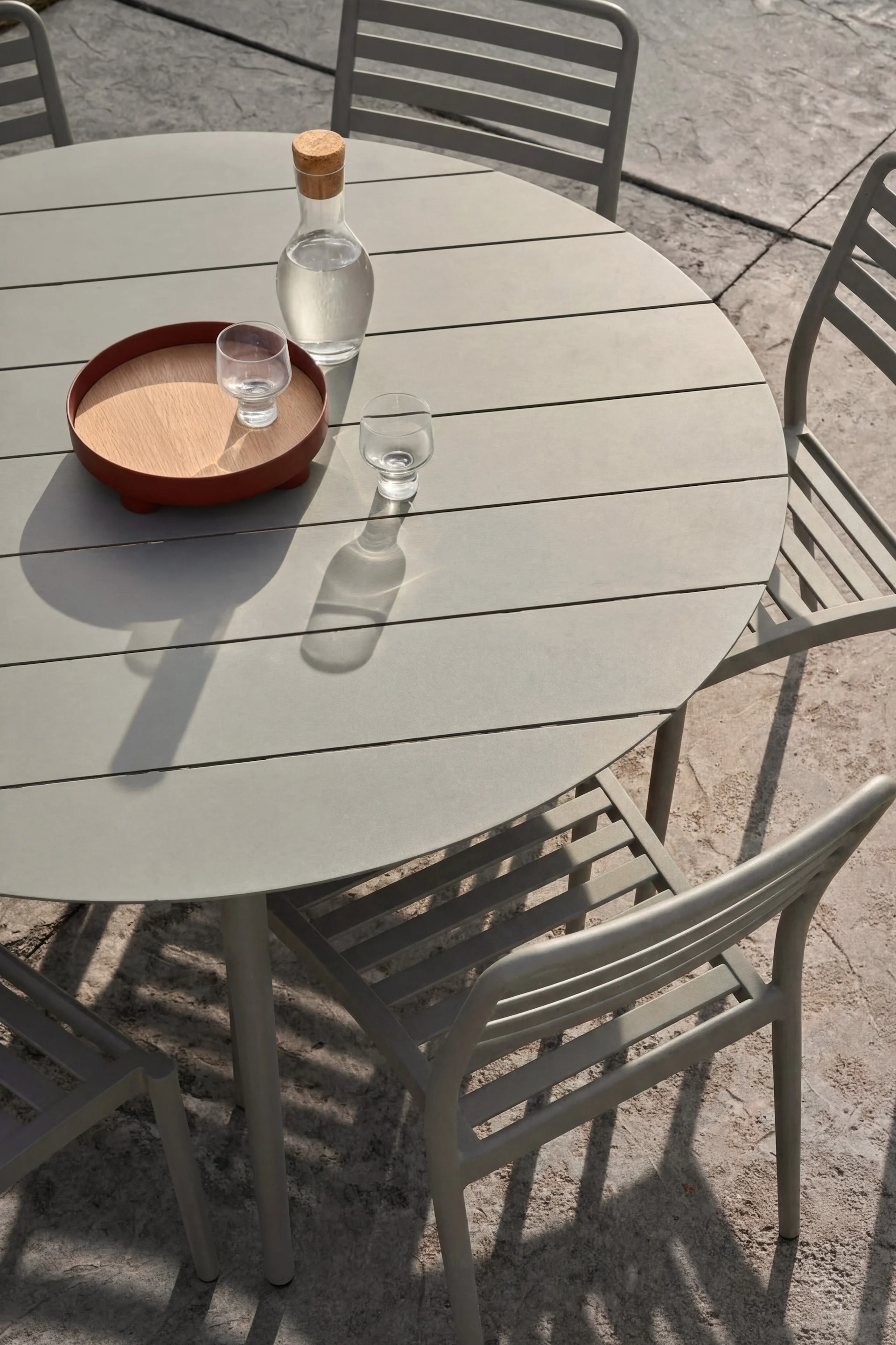 Runder grauer Gartentisch und drei Stühle. Glaskaraffe Wasser und zwei Gläser, eines auf Holztablett, auf dem Tisch.