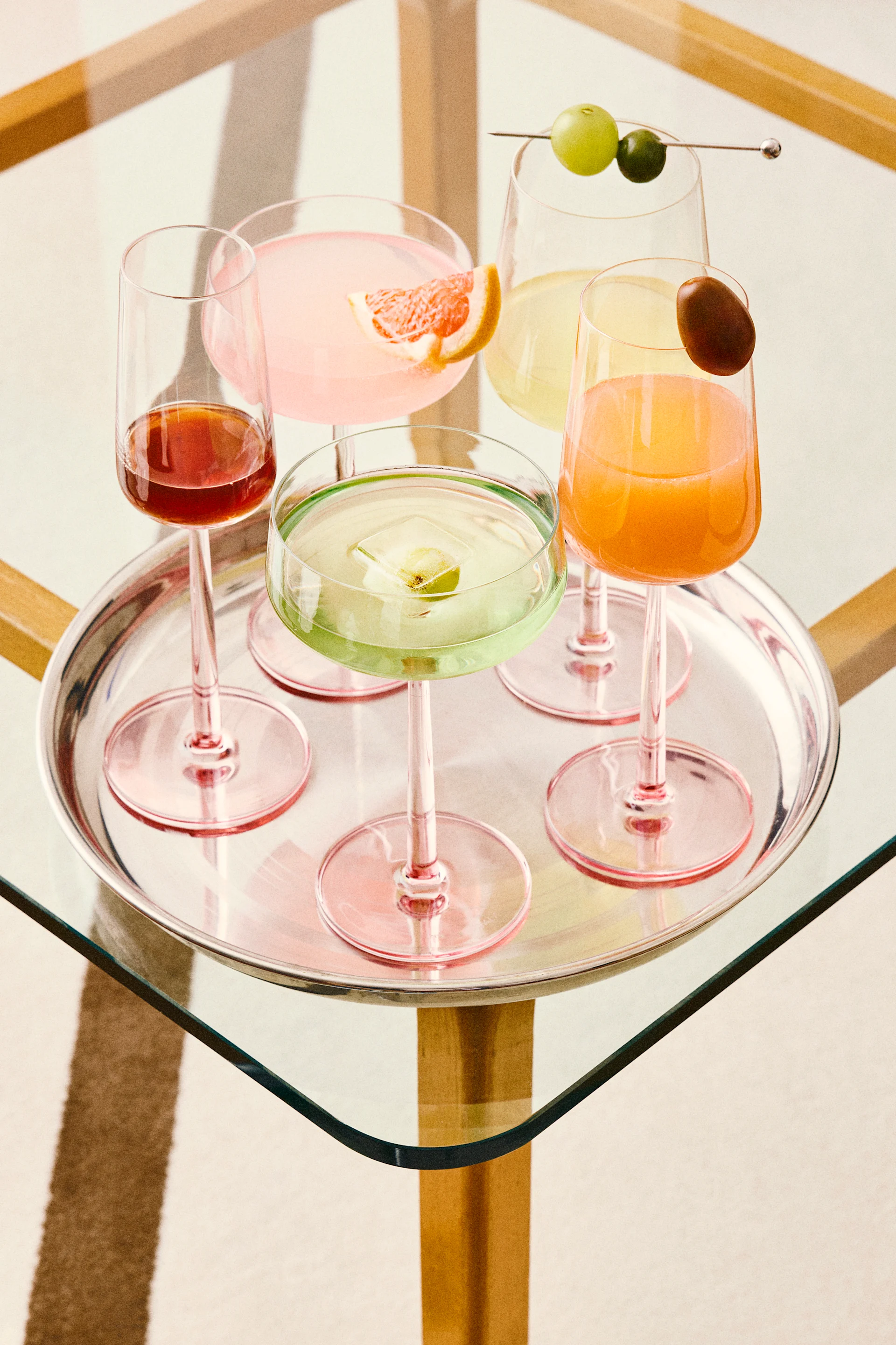 Fünf farbenfrohe Cocktails auf einem silbernen Tablett, darunter pinke, grüne, orange und rote Drinks mit verschiedenen Garnituren, auf einem Glastisch.