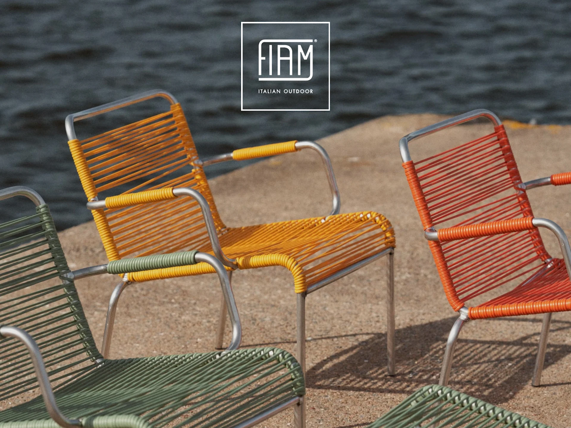 Drei farbenfrohe Outdoor-Stühle in Gelb, Orange und Grün mit geflochtenen Sitzen und Metallrahmen am Wasser.