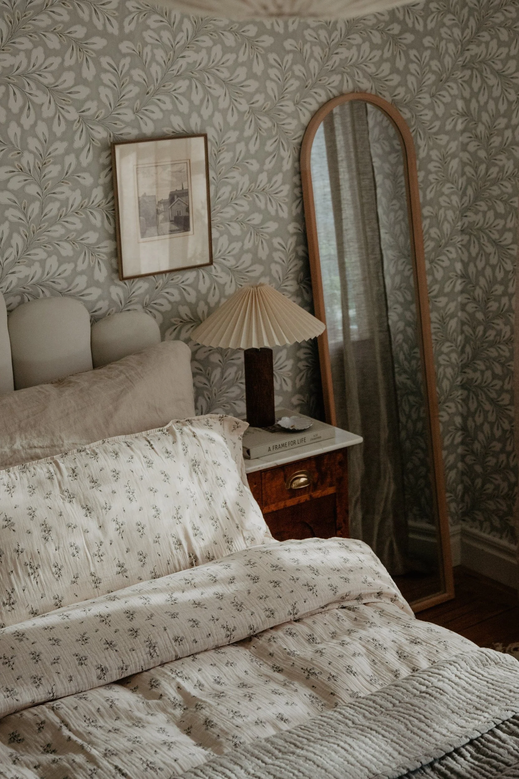 Ein Schlafzimmer mit cremefarbener gemusterter Bettwäsche, einer plissierten Lampe auf einem Holznachttisch und einem hohen Spiegel vor Blättertapete.