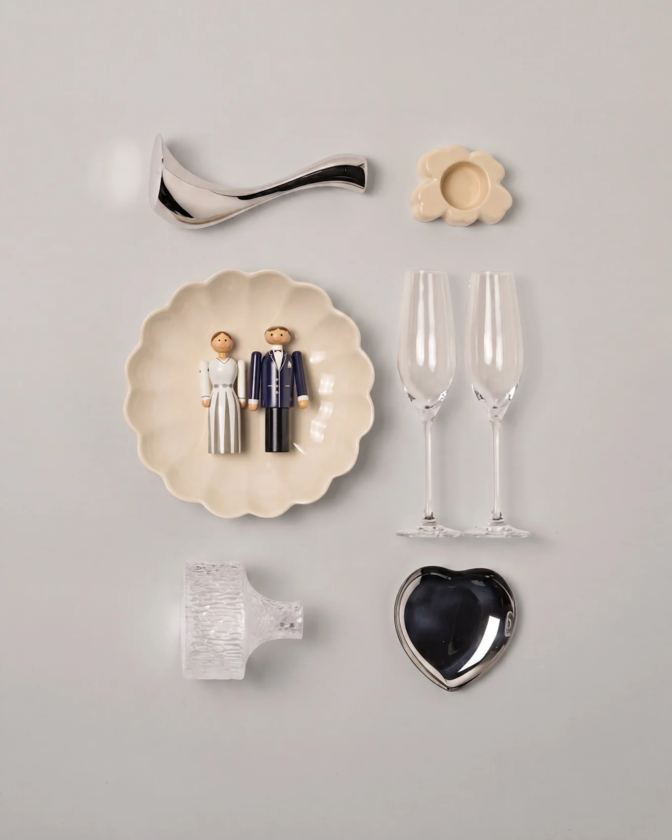 Hochzeits-Flat Lay mit hölzernen Braut- und Bräutigamfiguren auf einer Wellenschale, zwei Sektgläsern, einer silbernen Herzschale und verschiedenen Dekorationen.