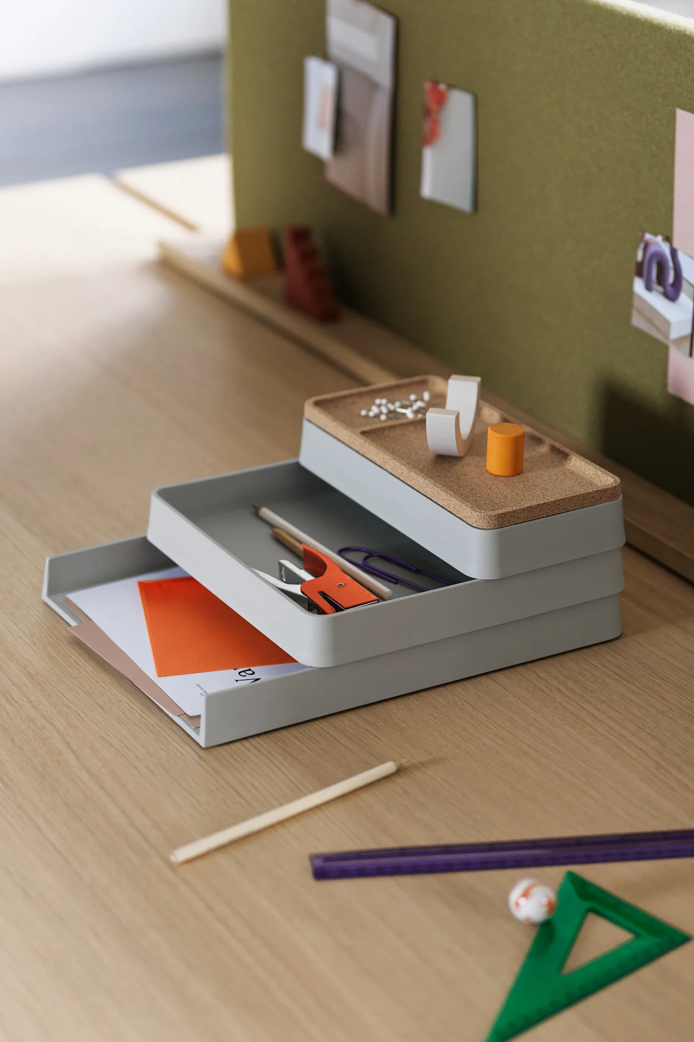Stapel von drei grauen Schreibtischorganisatoren mit korkgefütterter oberer Ablage, die Büromaterial wie Hefter, Nadeln und Papiere auf einem Holzschreibtisch enthält.