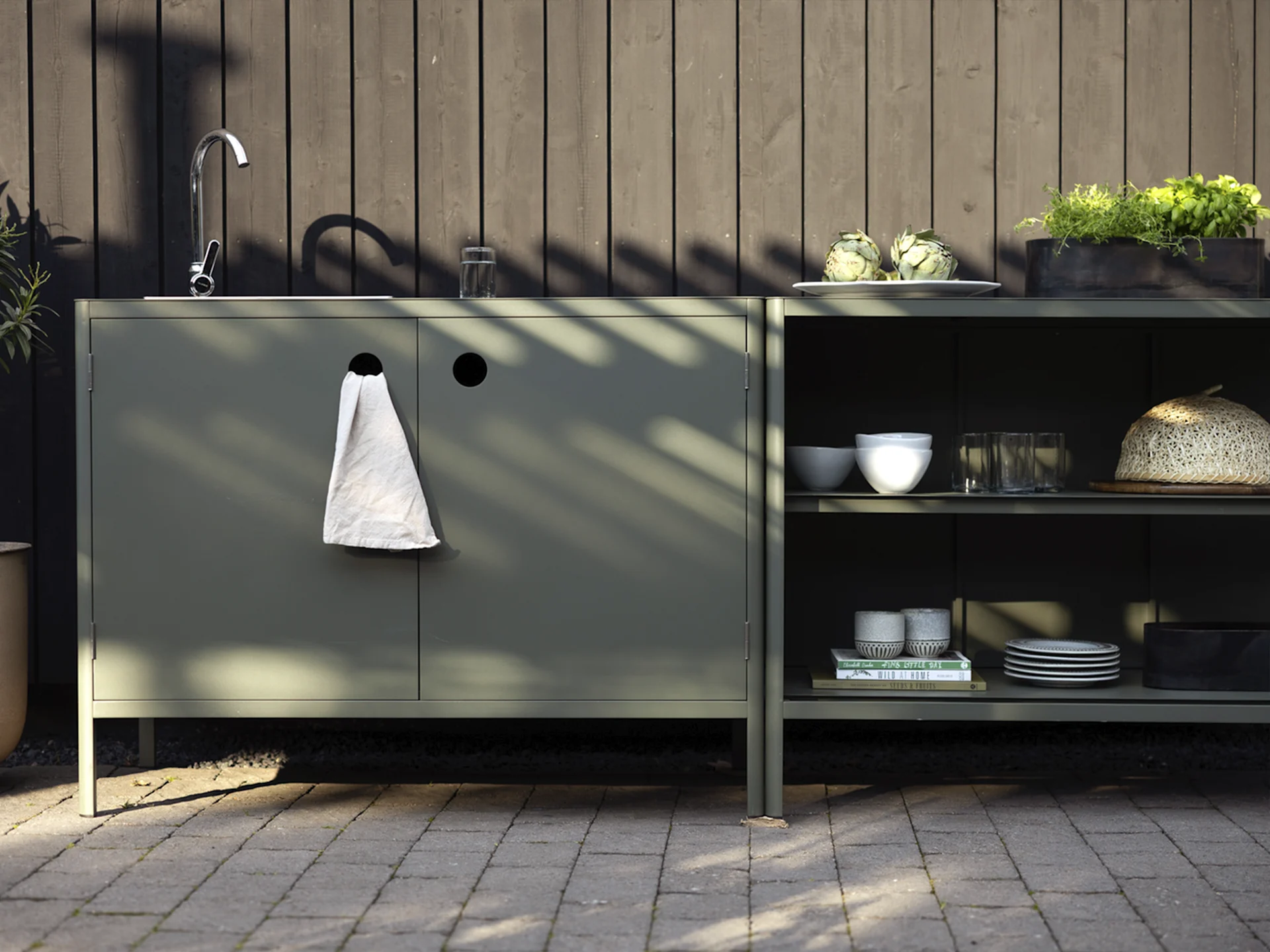 Außenküche mit dunkelgrünen Schränken, Spüle, Wasserhahn und Regalen mit Artischocken, Kräutern und Geschirr.