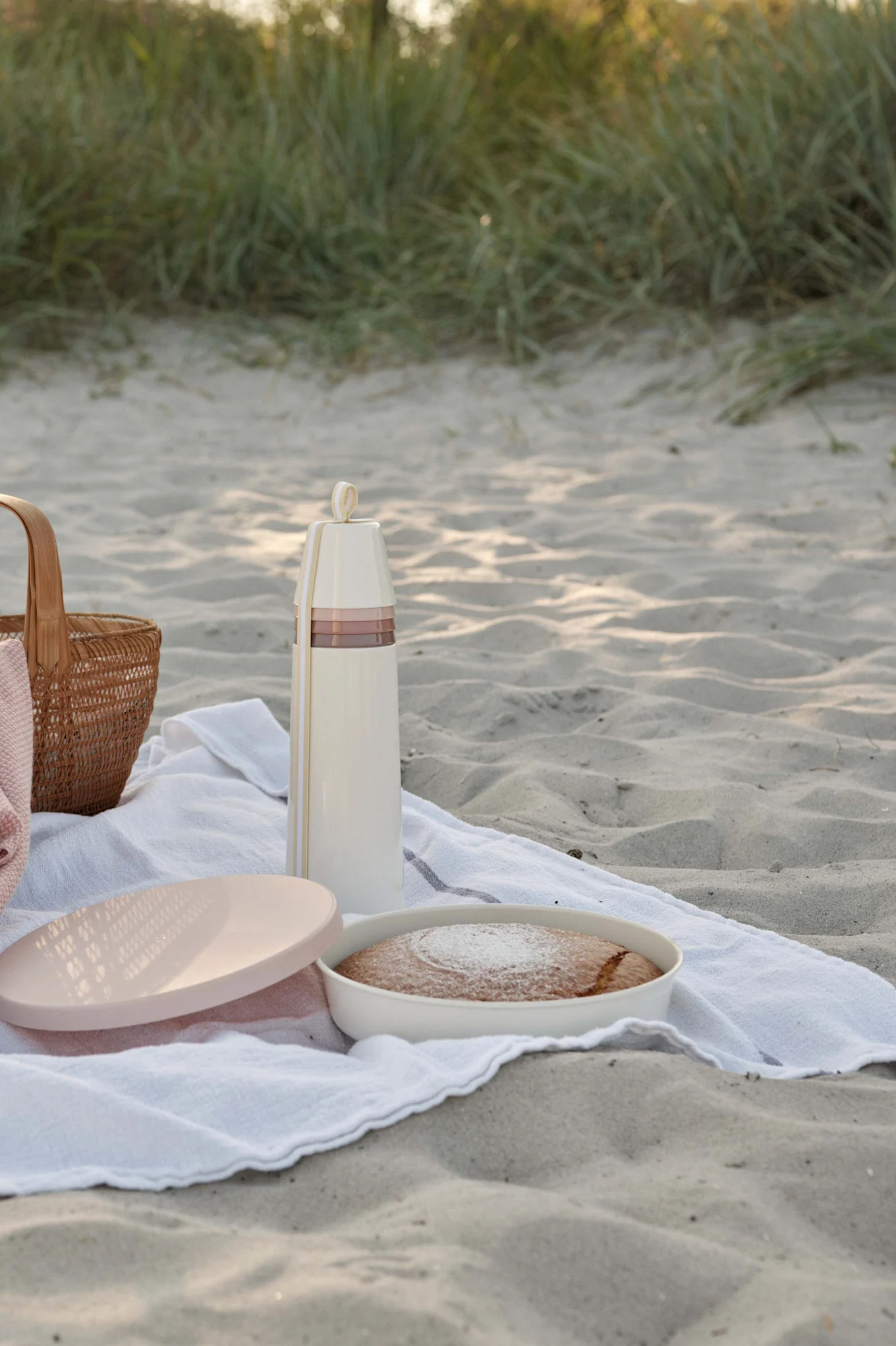 Picknick am Sandstrand mit cremefarbener Thermoskanne, Kuchen mit Puderzucker und geflochtenem Korb.