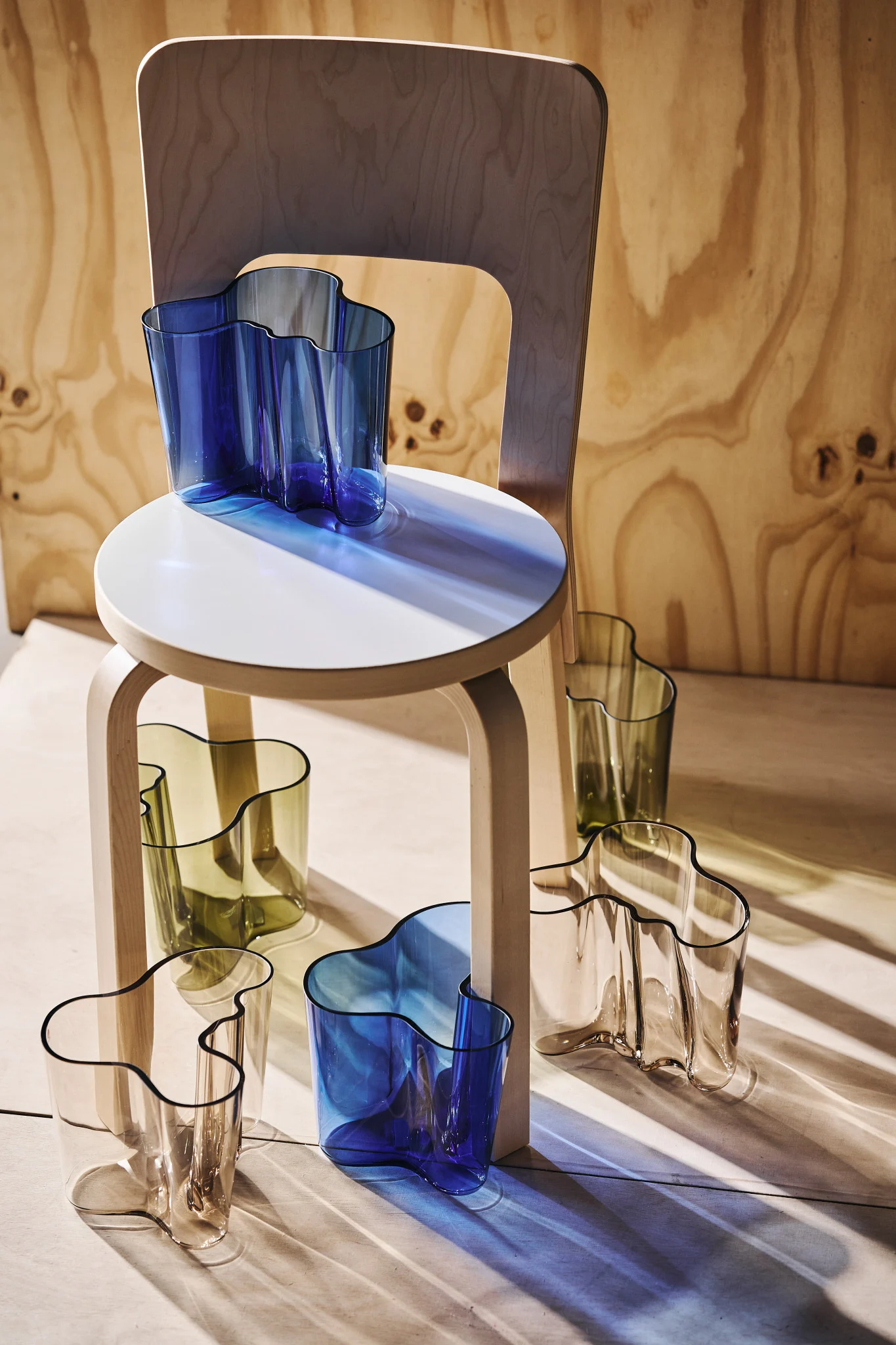 Mehrere wellenförmige Glasvasen in Blau, Olivgrün und Klar, auf und um einen hellen Holzschemel mit einem Holzstuhl im Hintergrund arrangiert. Sonnenlicht wirft farbige Schatten.