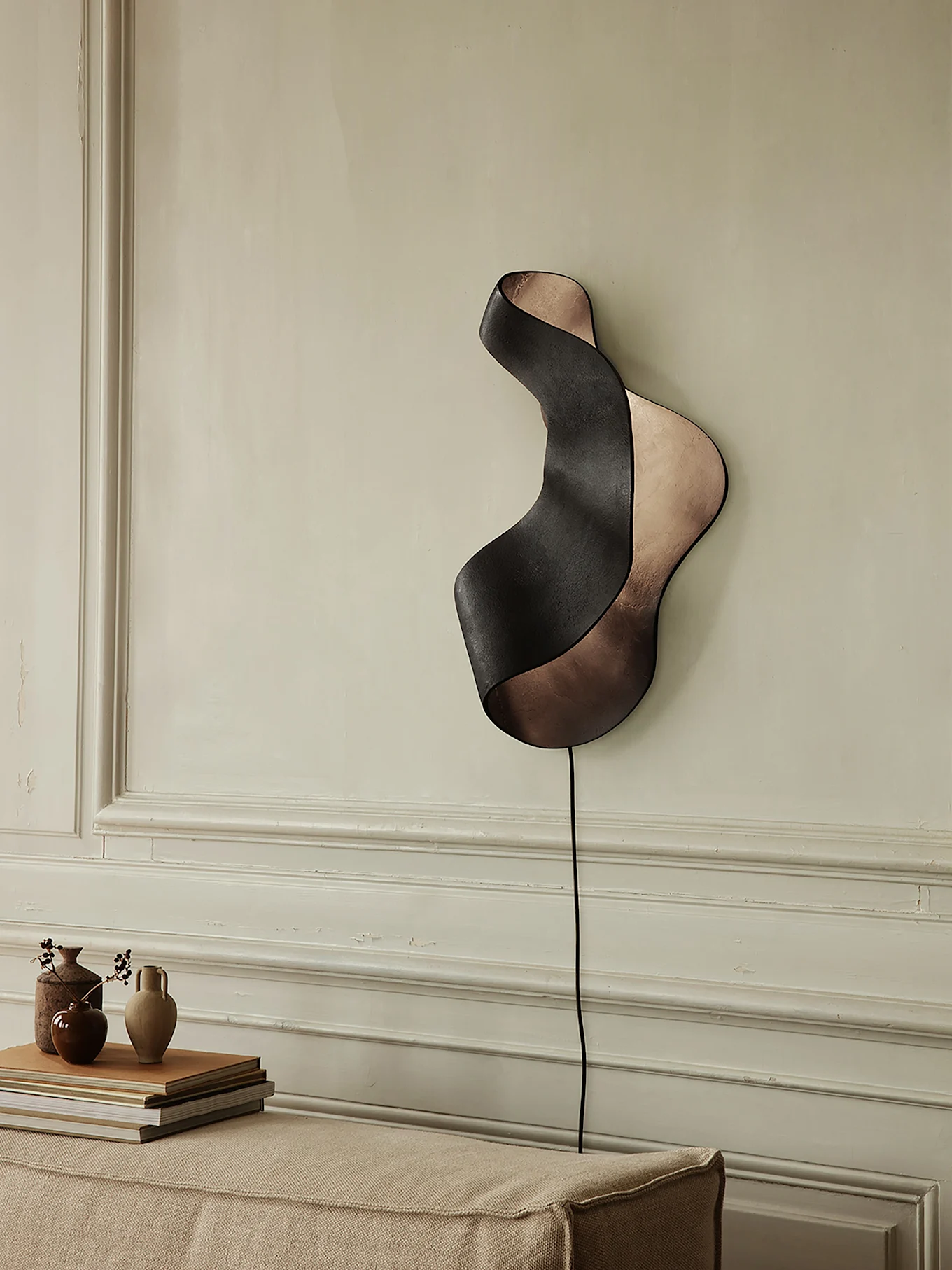 Schwarze und bronzefarbene wellenförmige Wandleuchte an heller Wand über Sofa mit Vasen und Büchern.