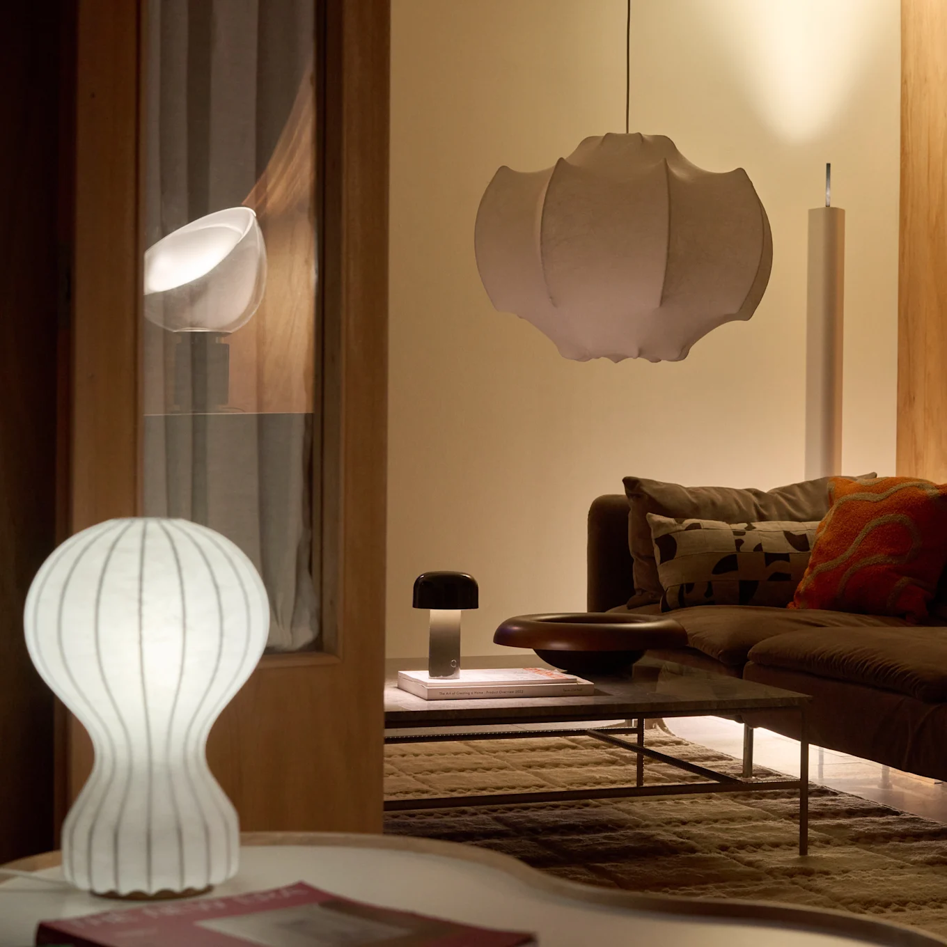 Ein Wohnzimmer mit einem braunen Sofa und Couchtisch, beleuchtet von einer weißen gerippten Tischlampe, einer dunklen Pilzlampe und einer großen plissierten Pendelleuchte, die ein warmes Licht erzeugt.
