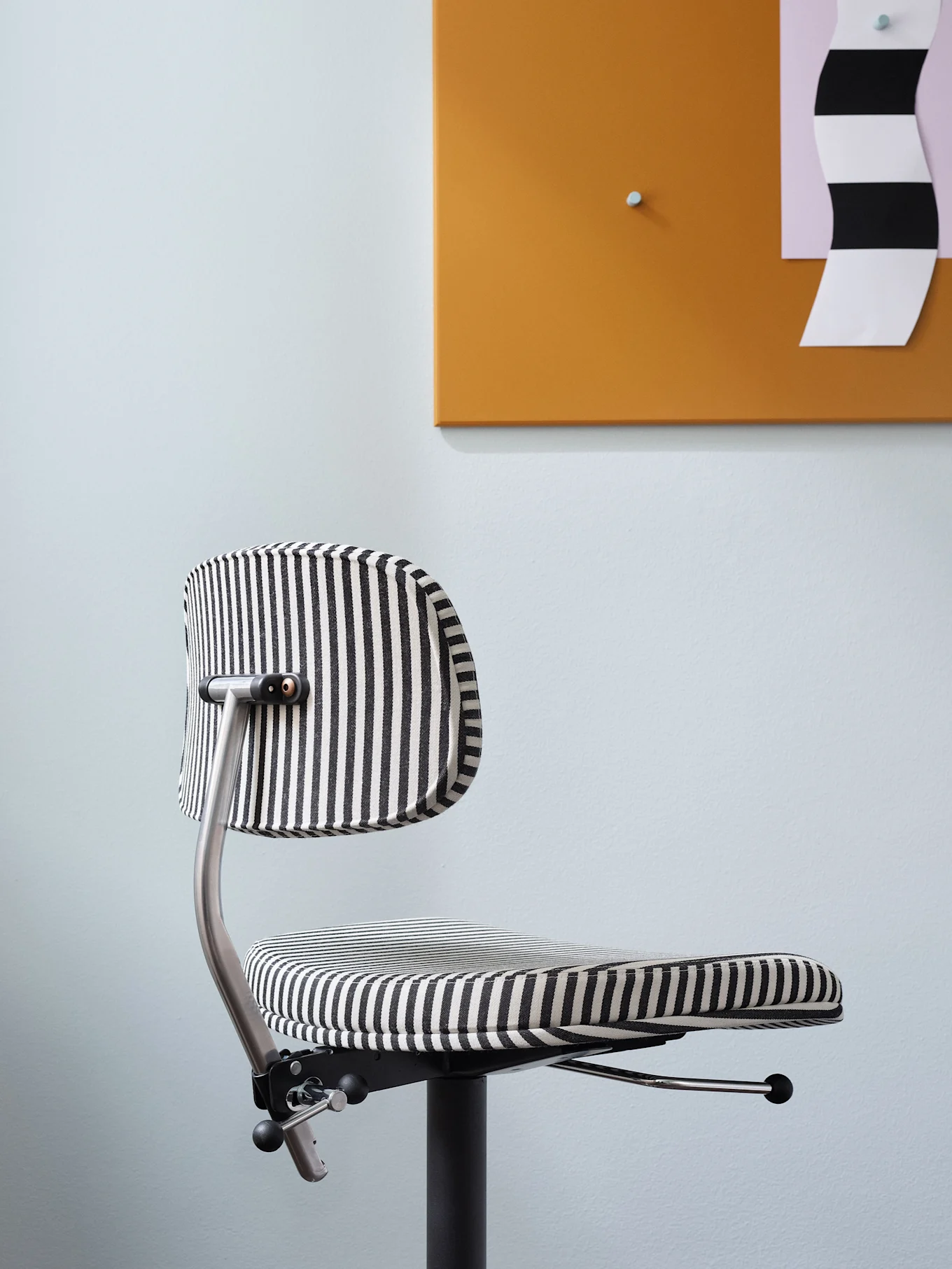 Ein moderner Bürostuhl mit schwarzen und weißen vertikalen Streifen vor einer hellblauen Wand und einer orangen Tafel.