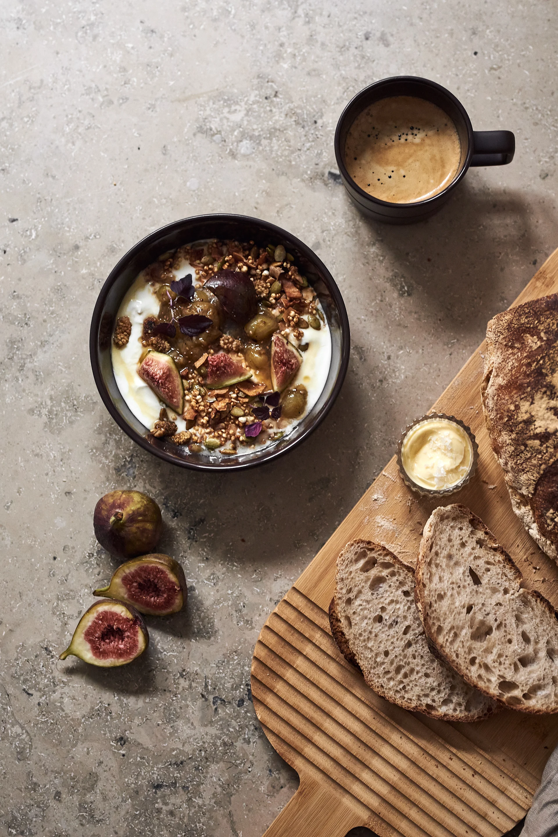 Die braune Schüssel aus der Lines-Serie von NJRD mit Müsli, einer Tasse Kaffee und einem Holzbrett mit geschnittenem Brot darauf.